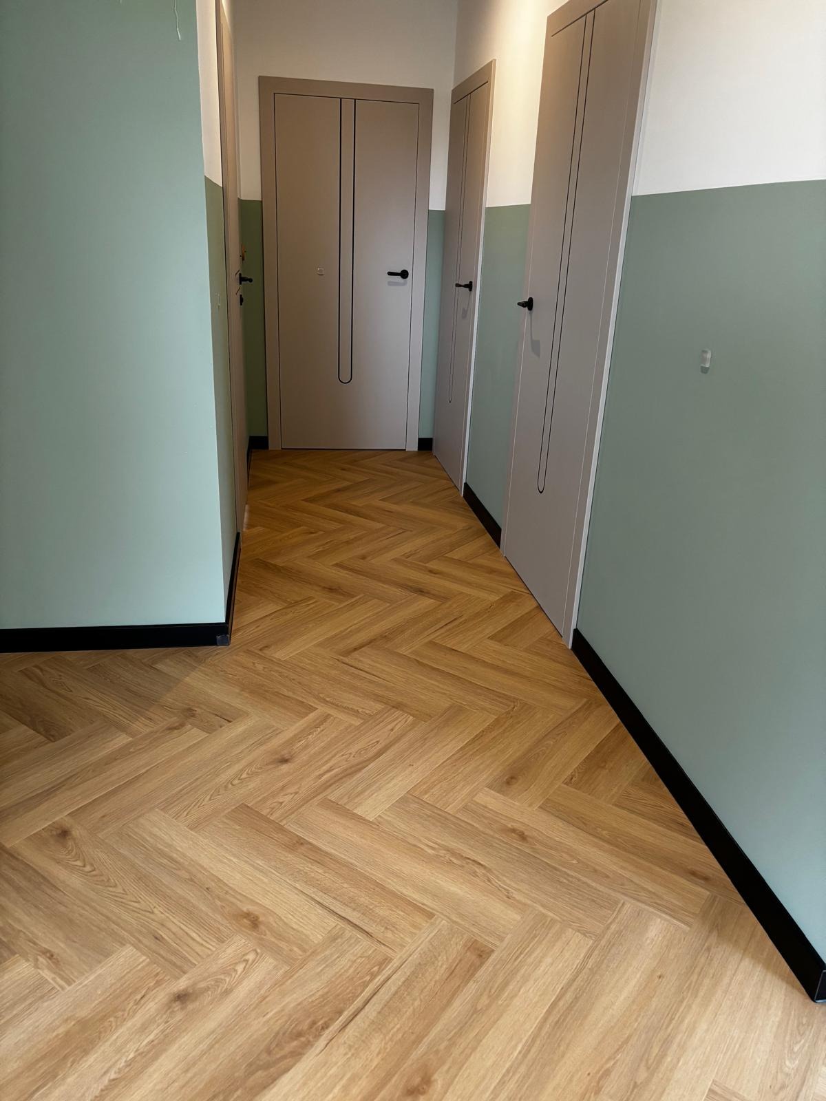Ułożona podłoga z paneli w jodełkę, widok na korytarz z trzema parami drzwi w kolorze beżowym, ściany w dwóch odcieniach zieleni i bieli.