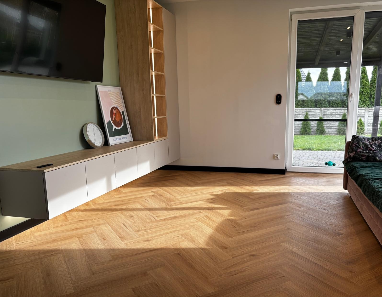 Jasne panele ułożone w jodełkę francuską w nowoczesnym salonie z minimalistyczną zabudową meblową i dużym oknem tarasowym z widokiem na ogród.