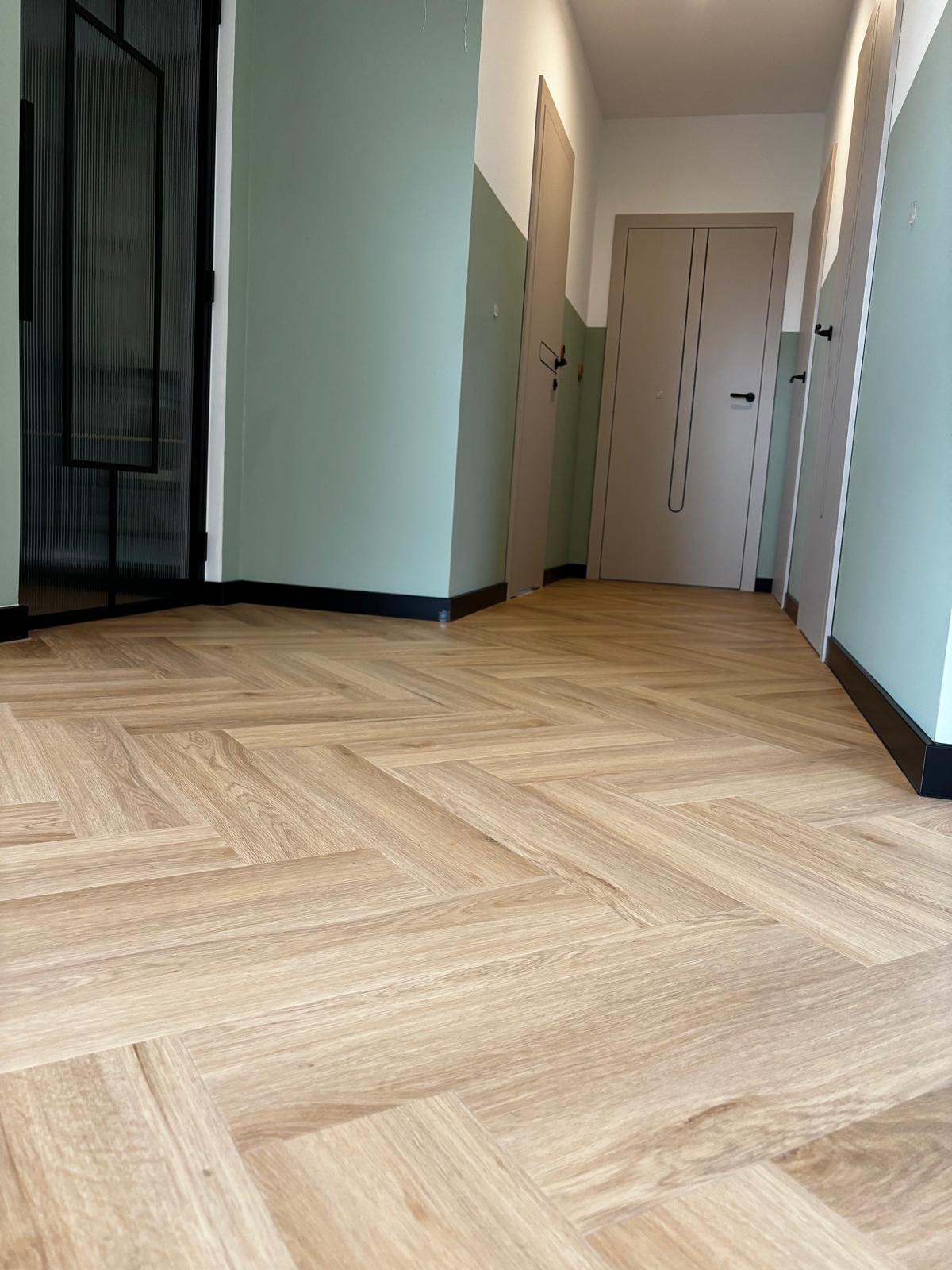 Jasne panele ułożone w jodełkę francuską w korytarzu z zielonymi ścianami i czarnymi listwami przypodłogowymi. Widoczne drzwi i przeszklenie w czarnej ramie.