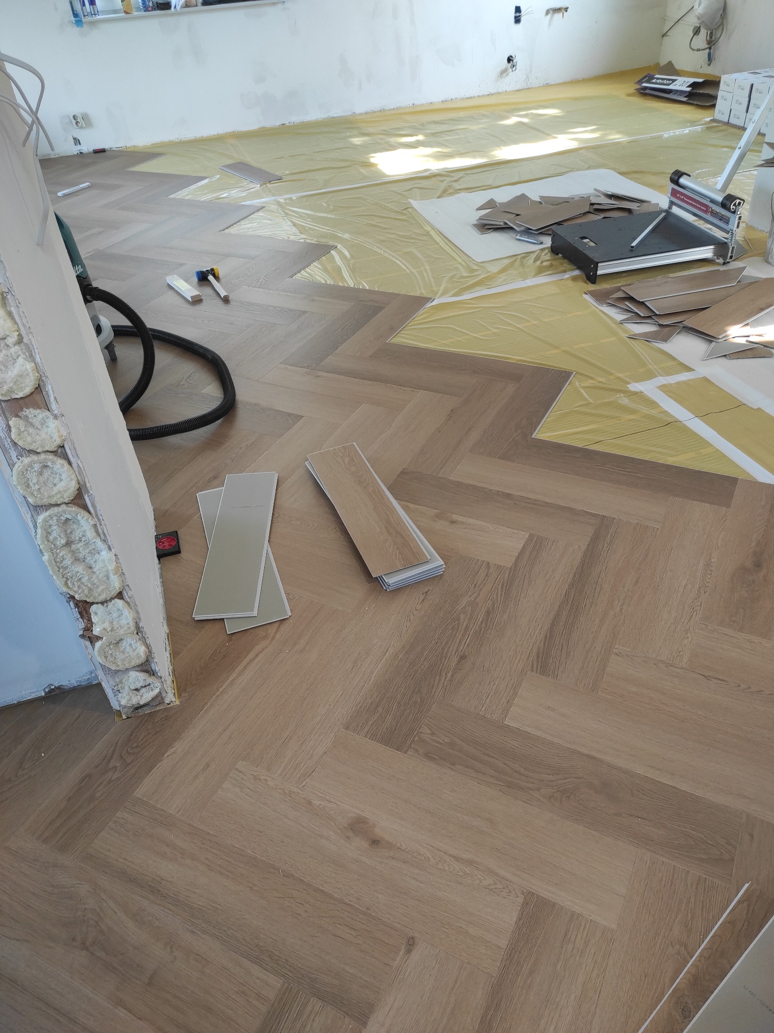 Układanie parkietu w jodełkę francuską. Widoczne narzędzia, folia ochronna i fragmenty paneli. Częściowo ułożona podłoga w jasnym odcieniu drewna.