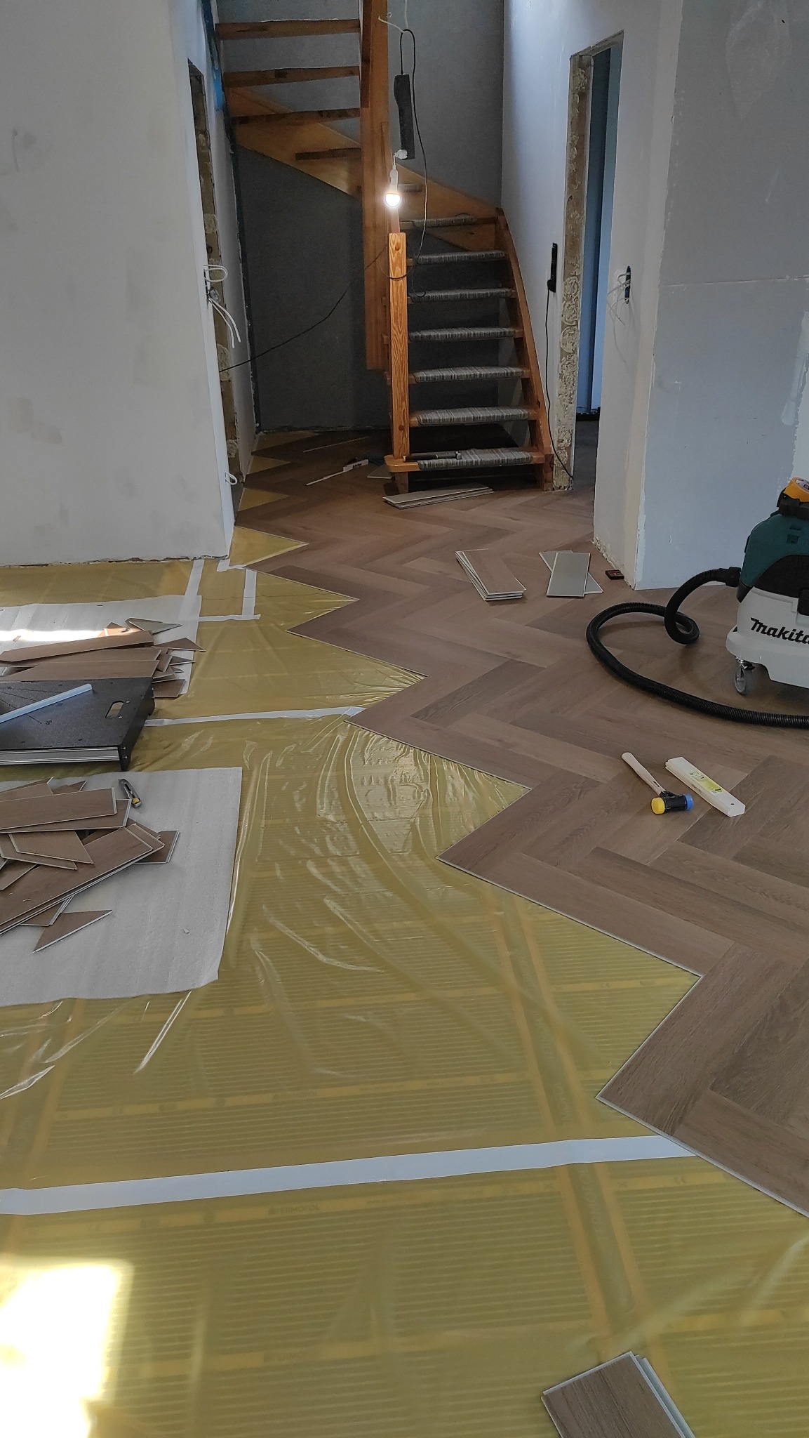 Układanie paneli w jodełkę na folii grzewczej, widoczne narzędzia i odkurzacz. Schody drewniane w tle. Etap prac wykończeniowych w nowym domu.