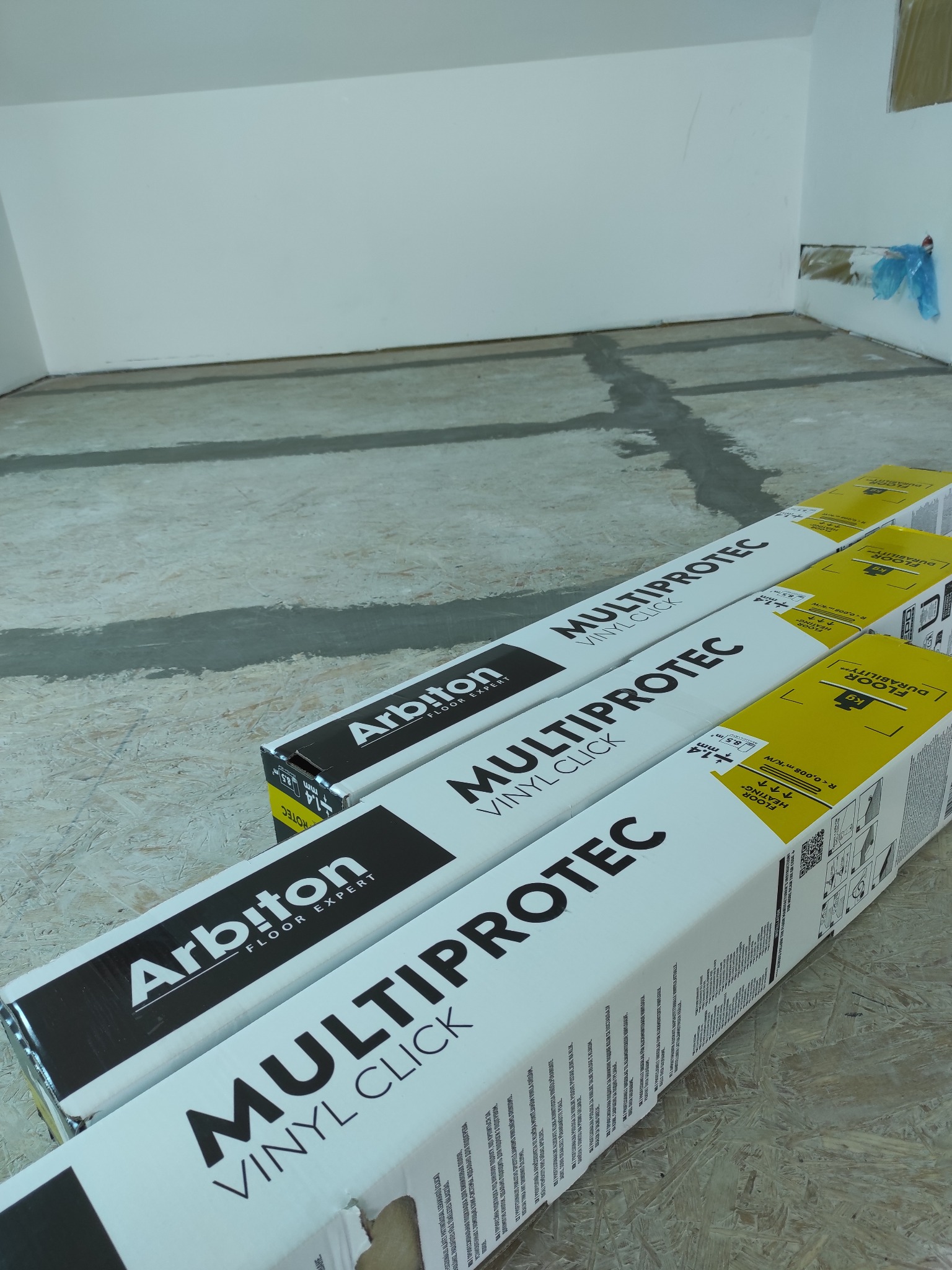 Przygotowanie do montażu paneli winylowych Arbiton Multiprotec. Widoczne paczki paneli na podłożu z widocznymi śladami po łatach. Białe ściany w tle.