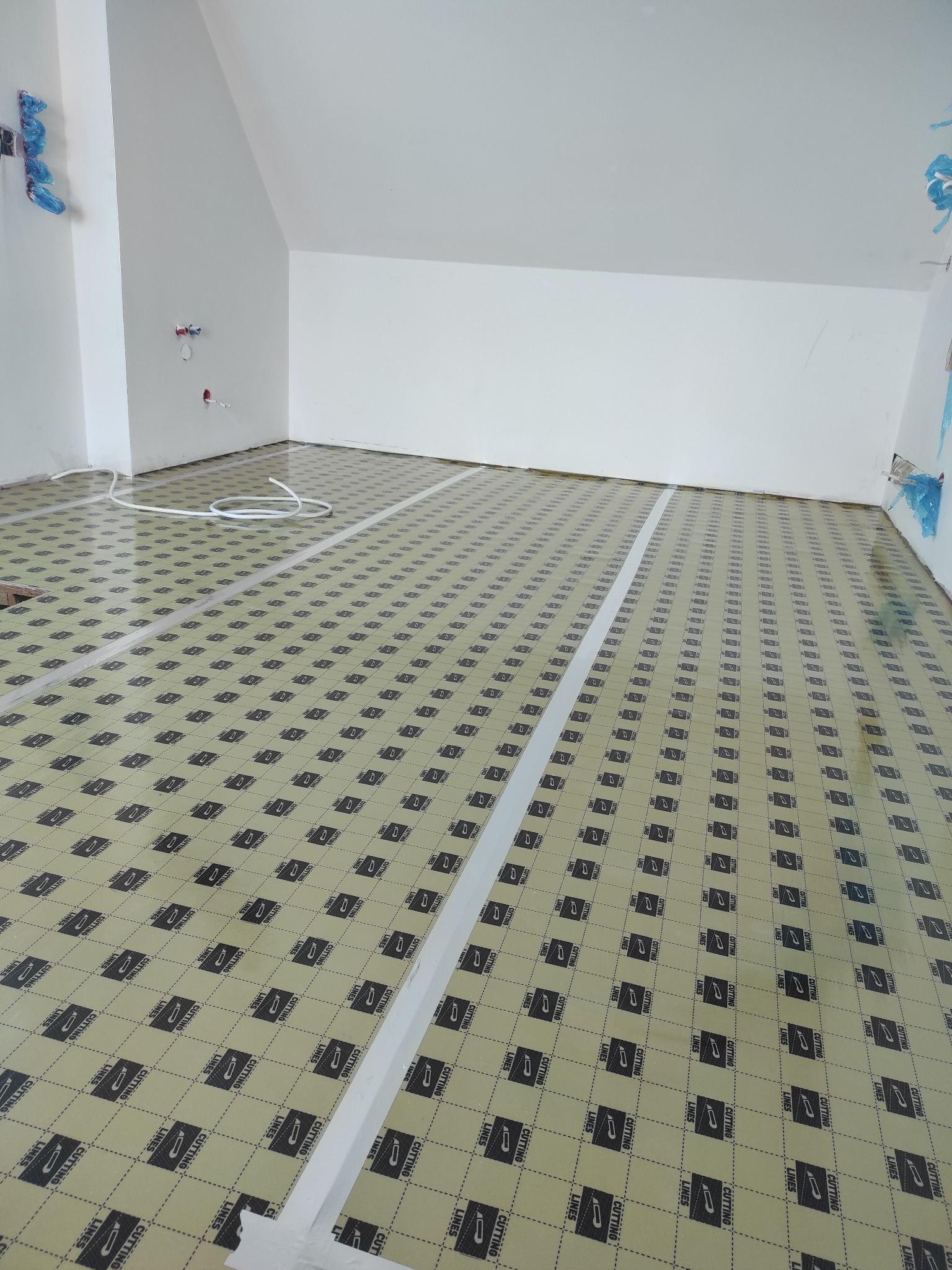 Przygotowana podłoga z folią izolacyjną i listwami dylatacyjnymi w jasnym pomieszczeniu ze skosami, gotowa do montażu paneli. Widok z góry.