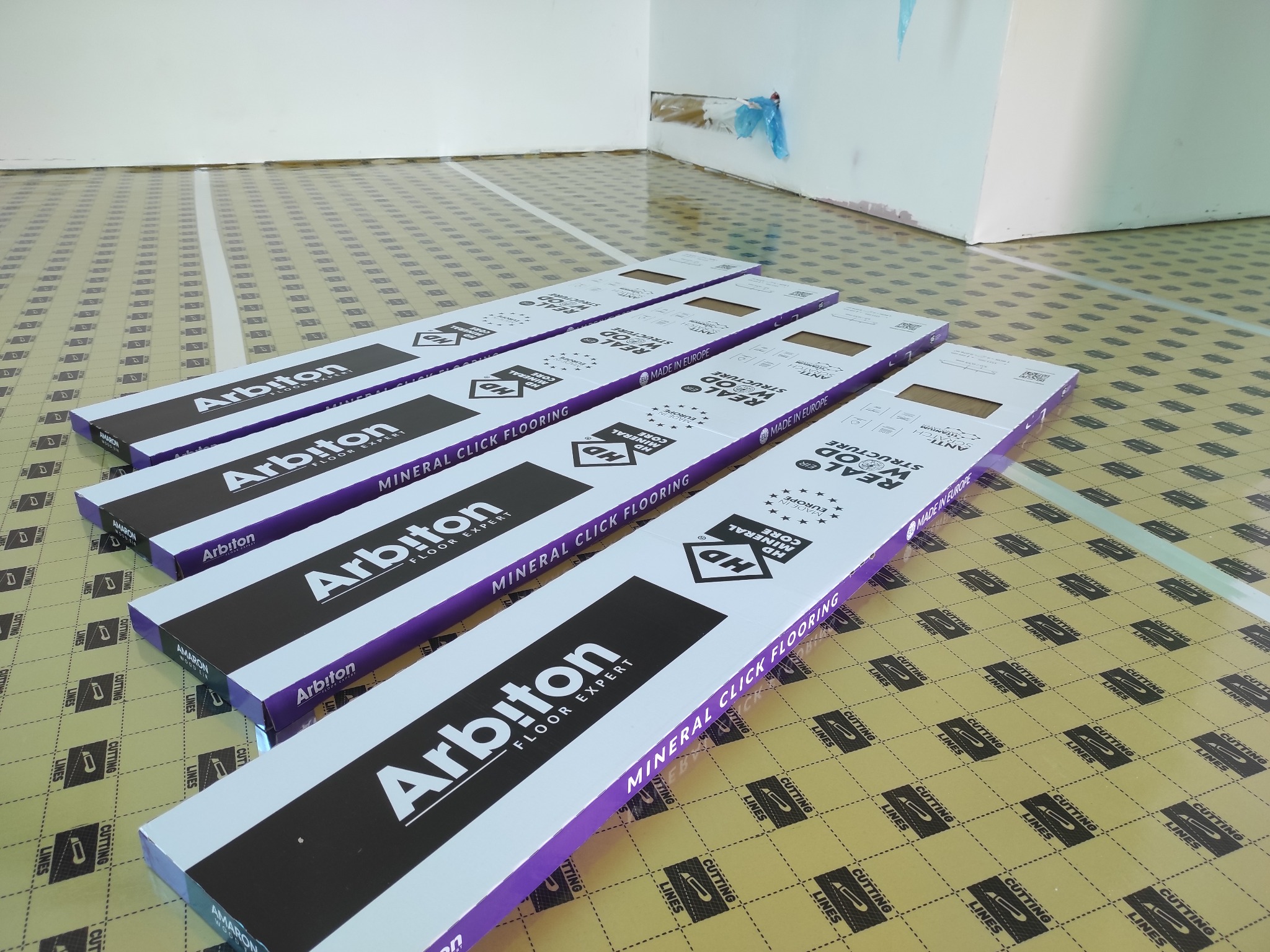 Paczki paneli podłogowych marki Arbiton leżące na podkładzie z nadrukiem linii cięcia. Widoczny fragment ściany w trakcie remontu.