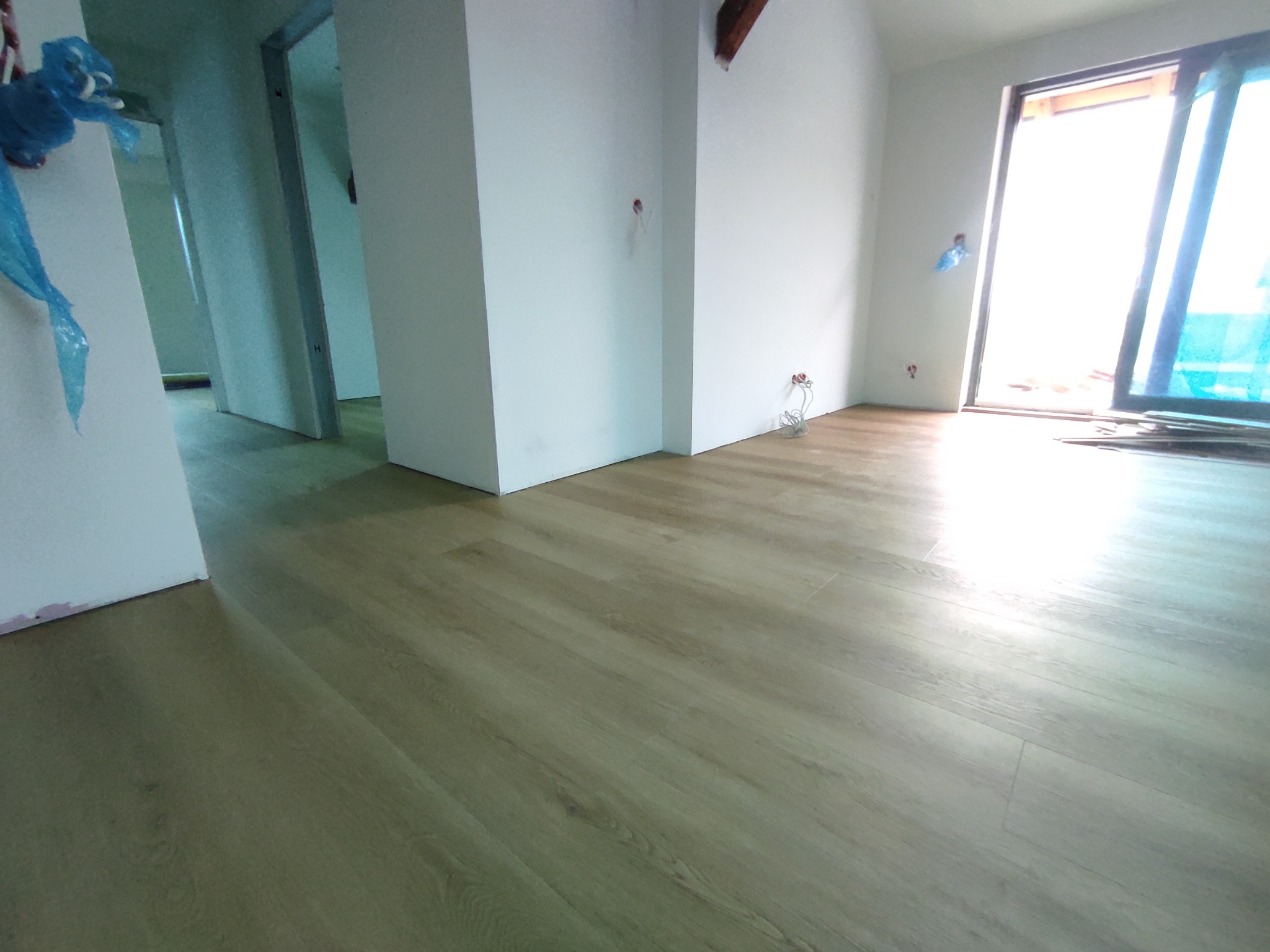 Jasne panele podłogowe ułożone w pustym, odnowionym pokoju z białymi ścianami i dużym oknem balkonowym. Widoczne ślady po instalacji elektrycznej.