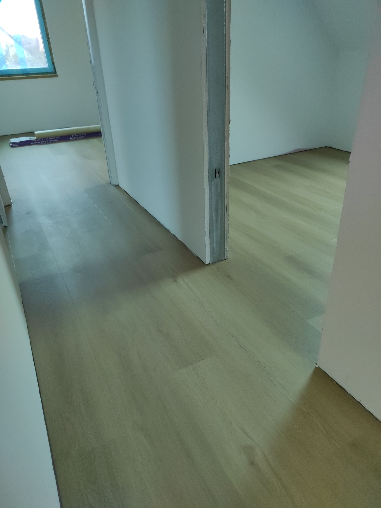 Jasne panele podłogowe ułożone w pokoju, widoczne przez otwarte drzwi. W tle opakowanie paneli przy ścianie. Ujęcie z niskiej perspektywy.