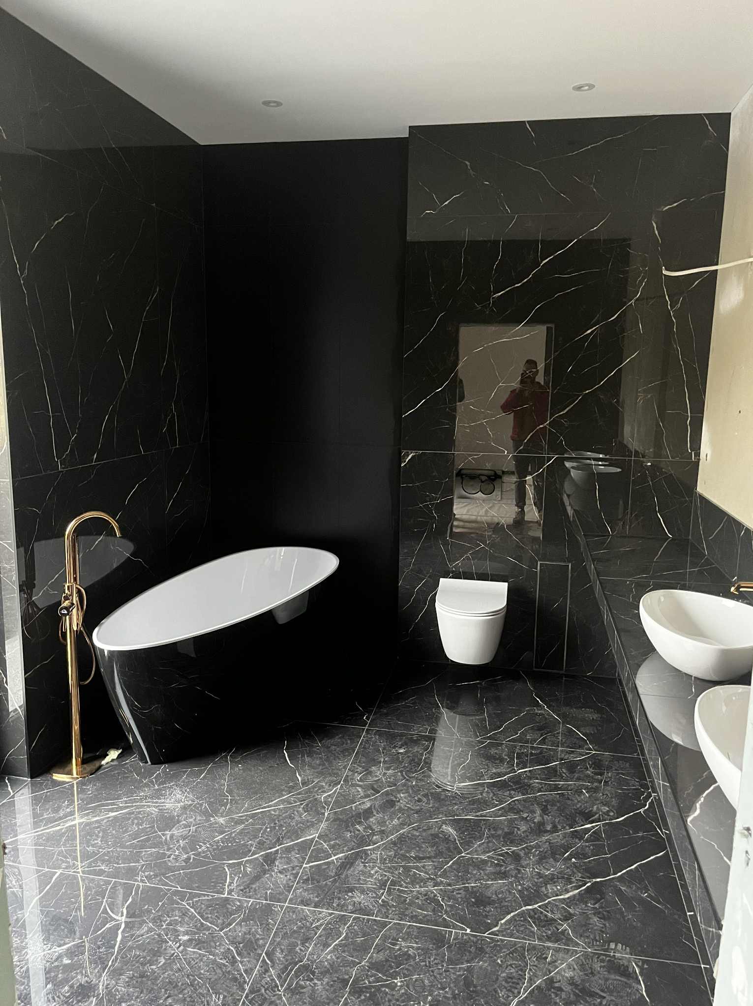 Elegancka łazienka w czerni i bieli z marmurowym wzorem, wolnostojąca wanna z złotą armaturą, umywalki nablatowe i toaleta. Widoczne odbicie osoby w lustrze.