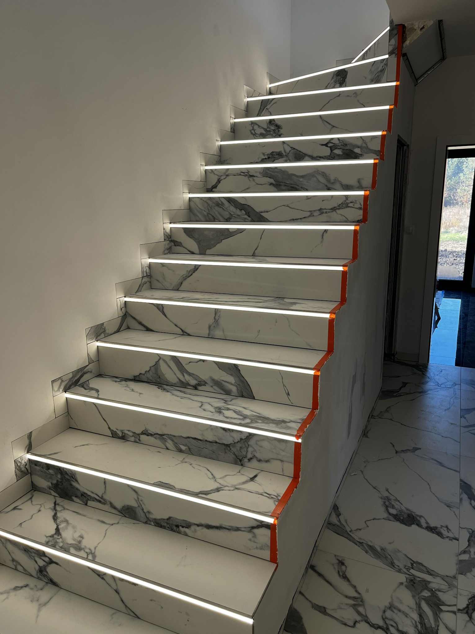 Marmurowe schody z podświetleniem LED, wykończone pomarańczową taśmą malarską, prowadzące do jasnego pomieszczenia z podłogą w tym samym wzorze.