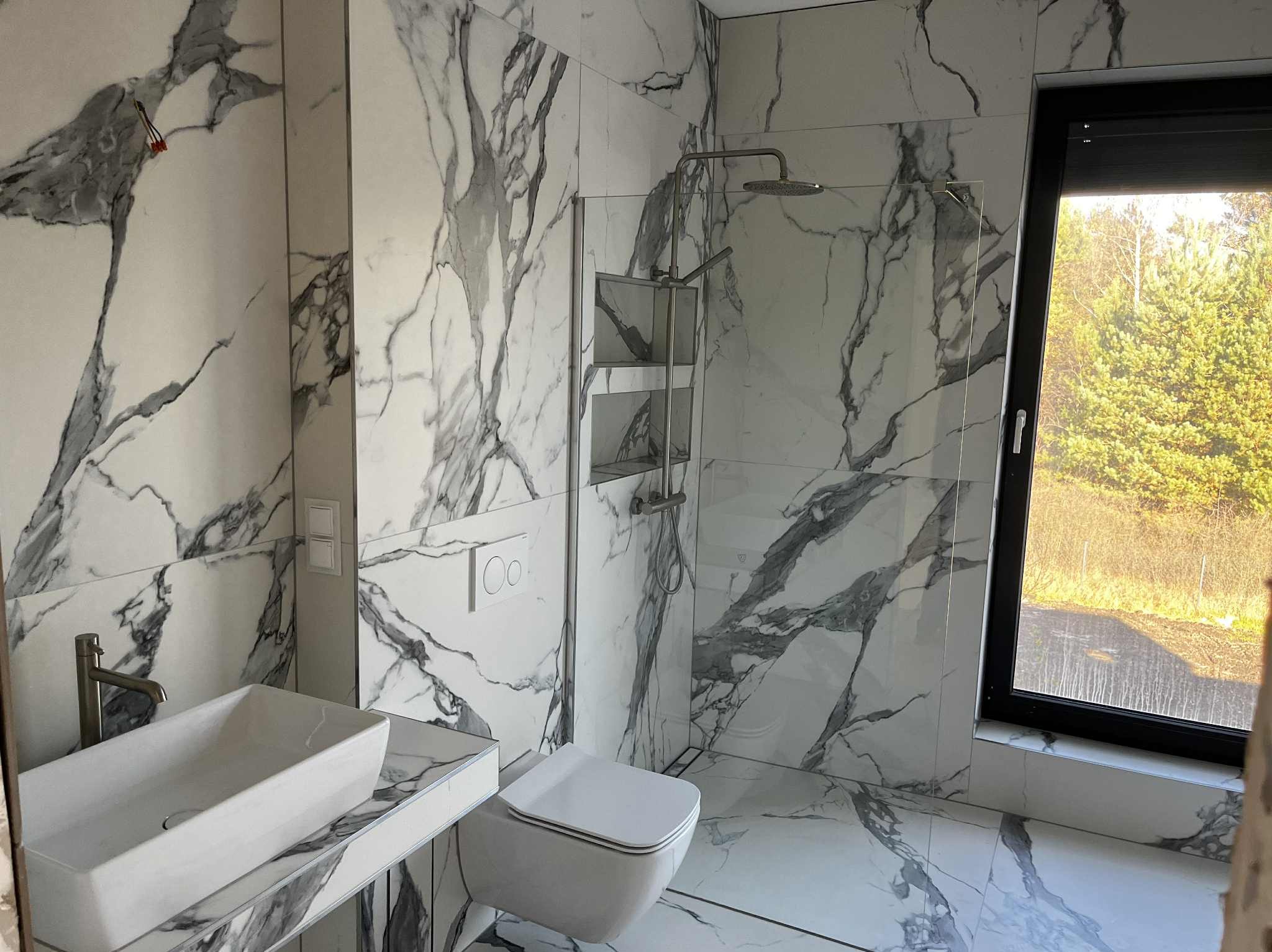 Elegancka łazienka z marmurowymi płytkami, kabiną prysznicową typu walk-in, umywalką nablatową i toaletą. Widok na zieleń za oknem.