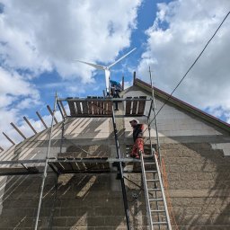 EkoSprzęt Filip Grygo - Montaż turbiny wiatrowej na dachu domu jednorodzinnego. Dwóch monterów na rusztowaniu, widok z dołu na tle błękitnego nieba z chmurami.