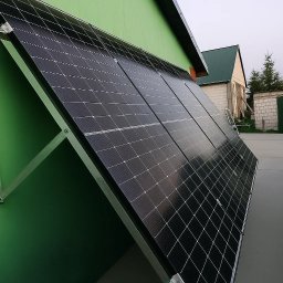 EkoSprzęt Filip Grygo - Ścienne panele słoneczne na zielonej elewacji budynku gospodarczego, z widocznym fragmentem dachu i zabudowań w tle, w pochmurny dzień. Montaż paneli pod kątem.