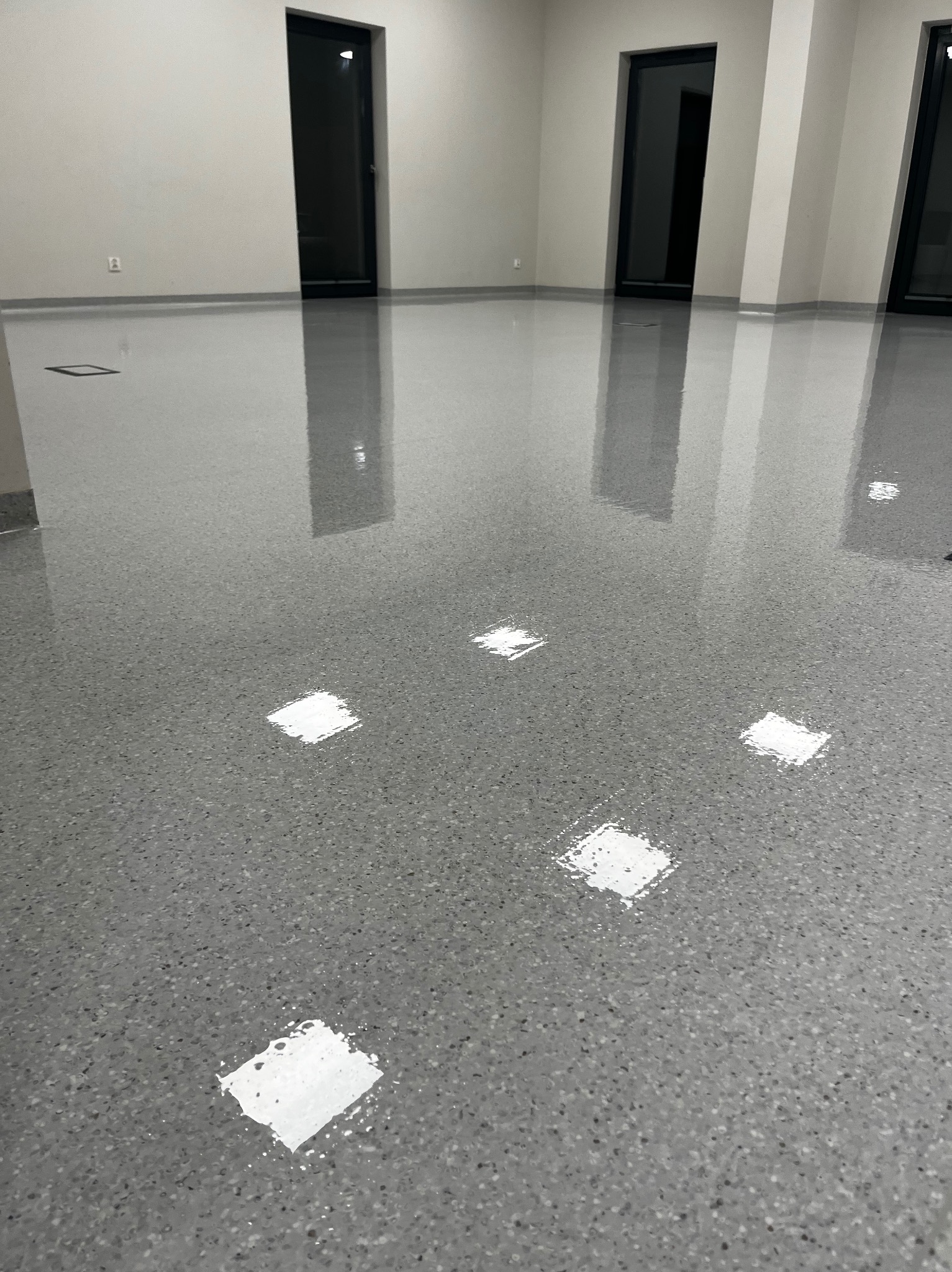 Błyszcząca, szara posadzka żywiczna w pustym, jasnym pomieszczeniu z trzema parami czarnych drzwi. Widoczne odbicia światła na gładkiej powierzchni.