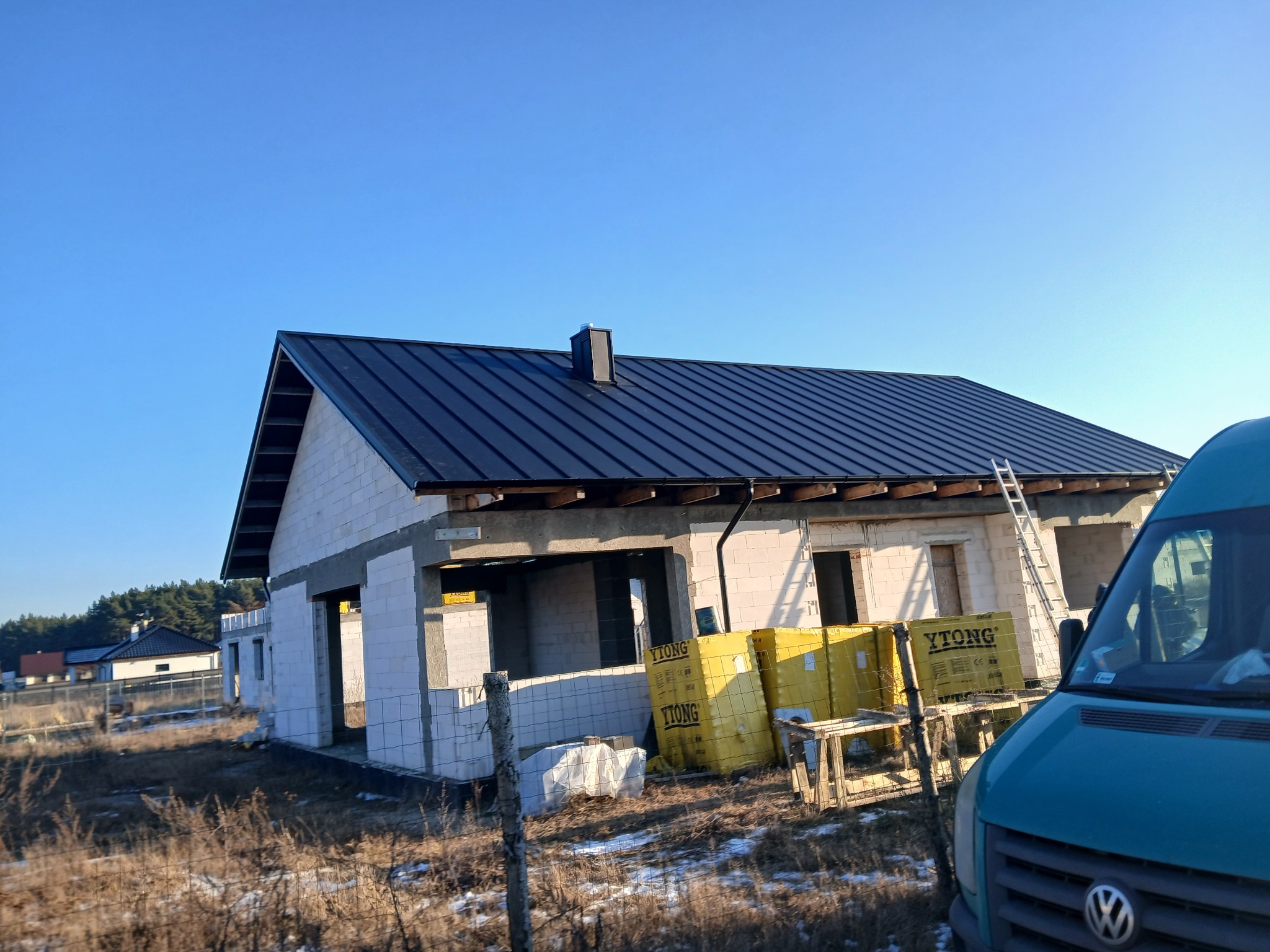 Dom w trakcie budowy z ciemnym dachem z blachy na tle błękitnego nieba. Widoczne palety z bloczkami Ytong i drabina oparta o ścianę.