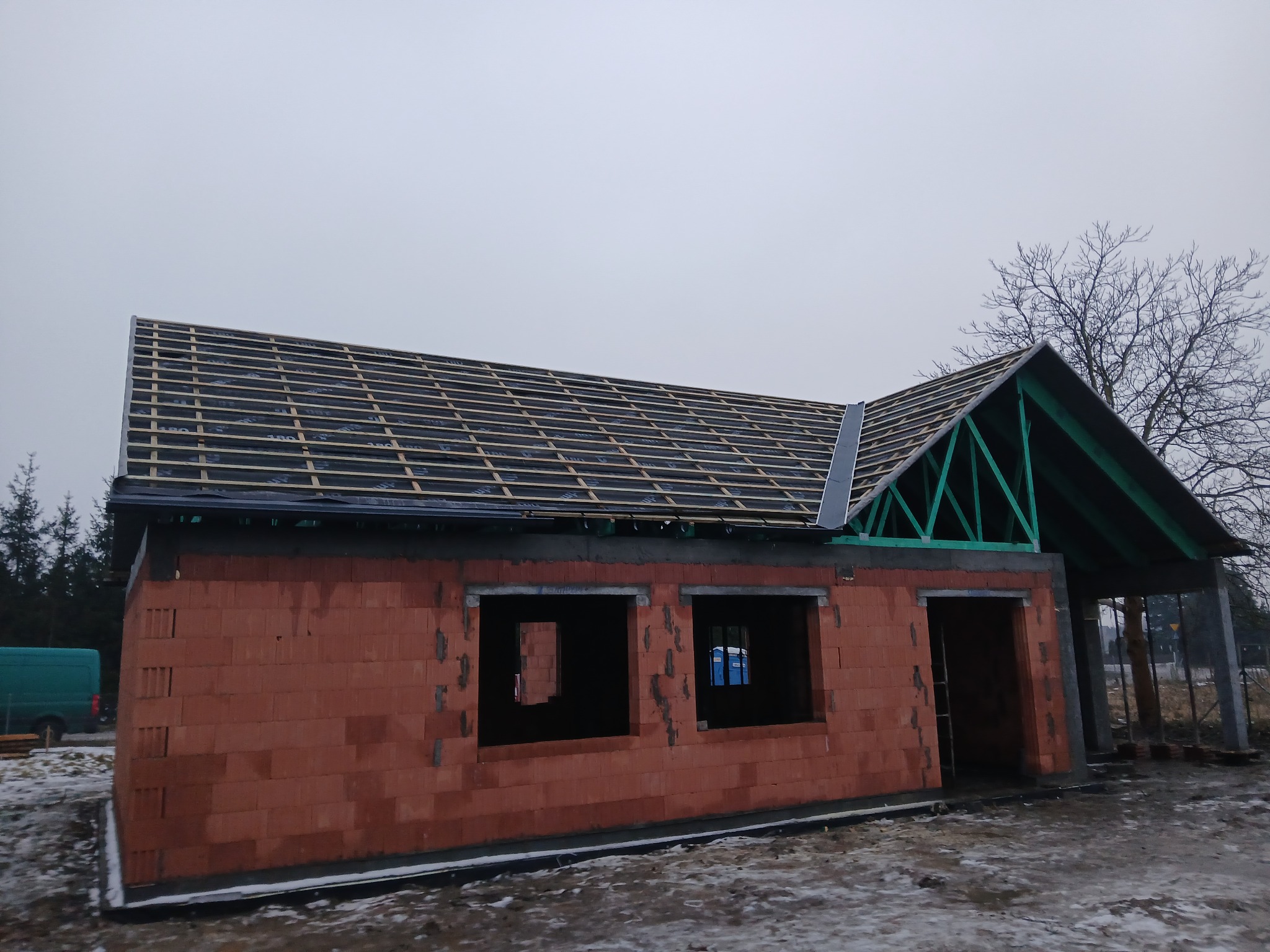 Budowa domu jednorodzinnego w stanie surowym otwartym: czerwone ściany z pustaków, więźba dachowa z łatami i folią, widoczne otwory okienne i drzwiowe.