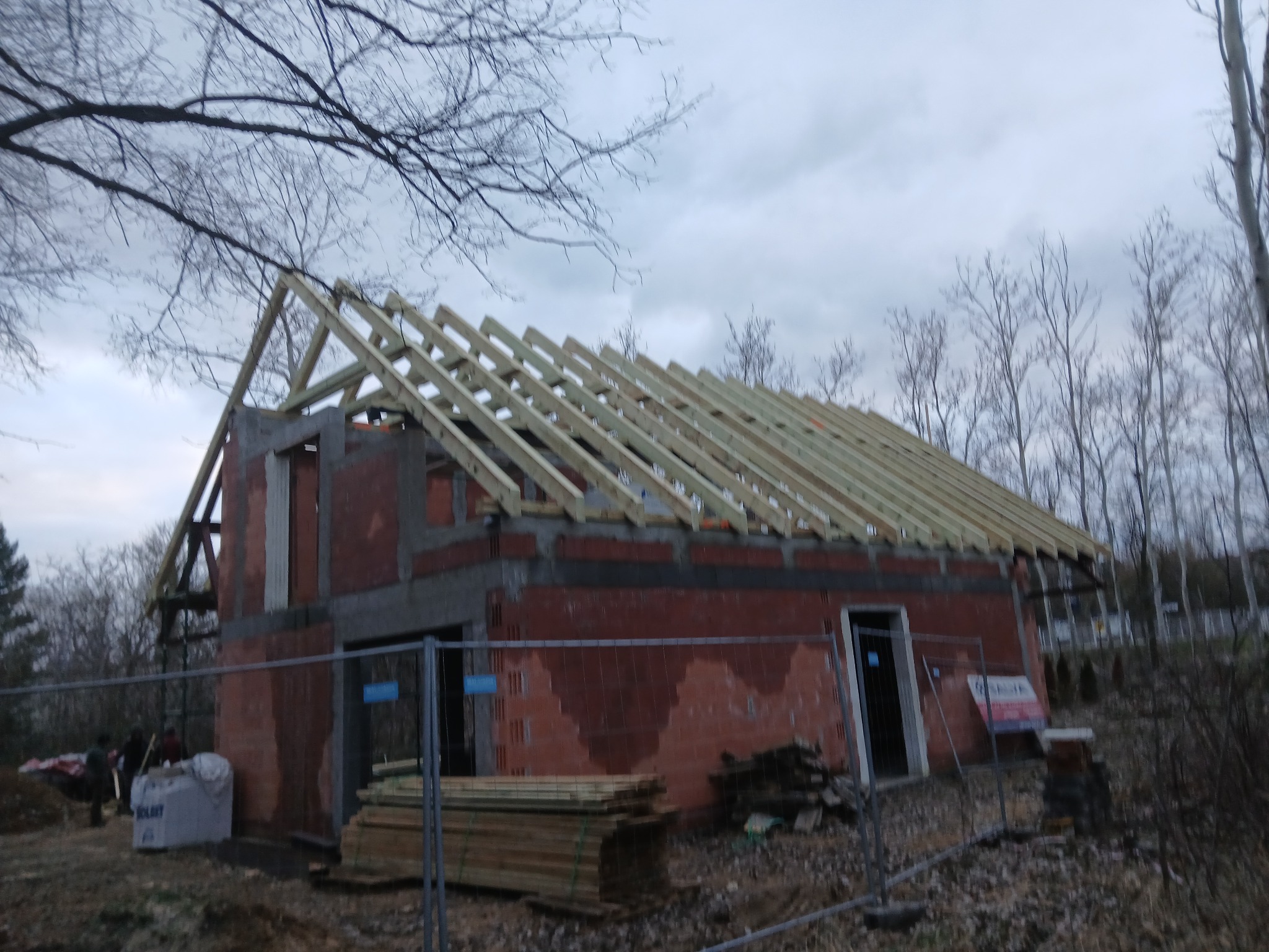 Wznoszenie więźby dachowej na murowanym budynku w stanie surowym otwartym, widoczne elementy konstrukcyjne i materiały budowlane na działce.