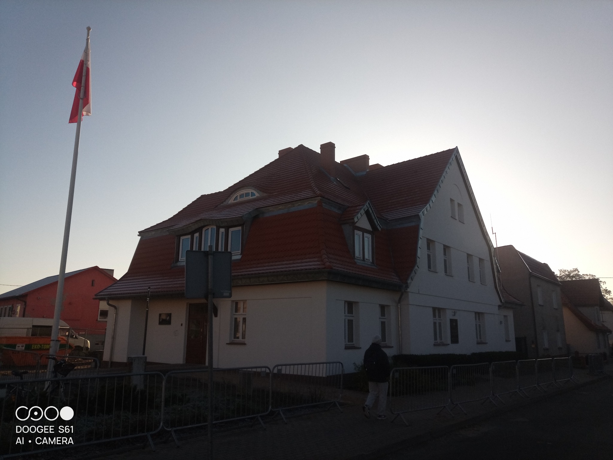 Budynek z czerwonym dachem w promieniach zachodzącego słońca, flaga Polski na maszcie. Widoczny fragment ulicy i ogrodzenie.