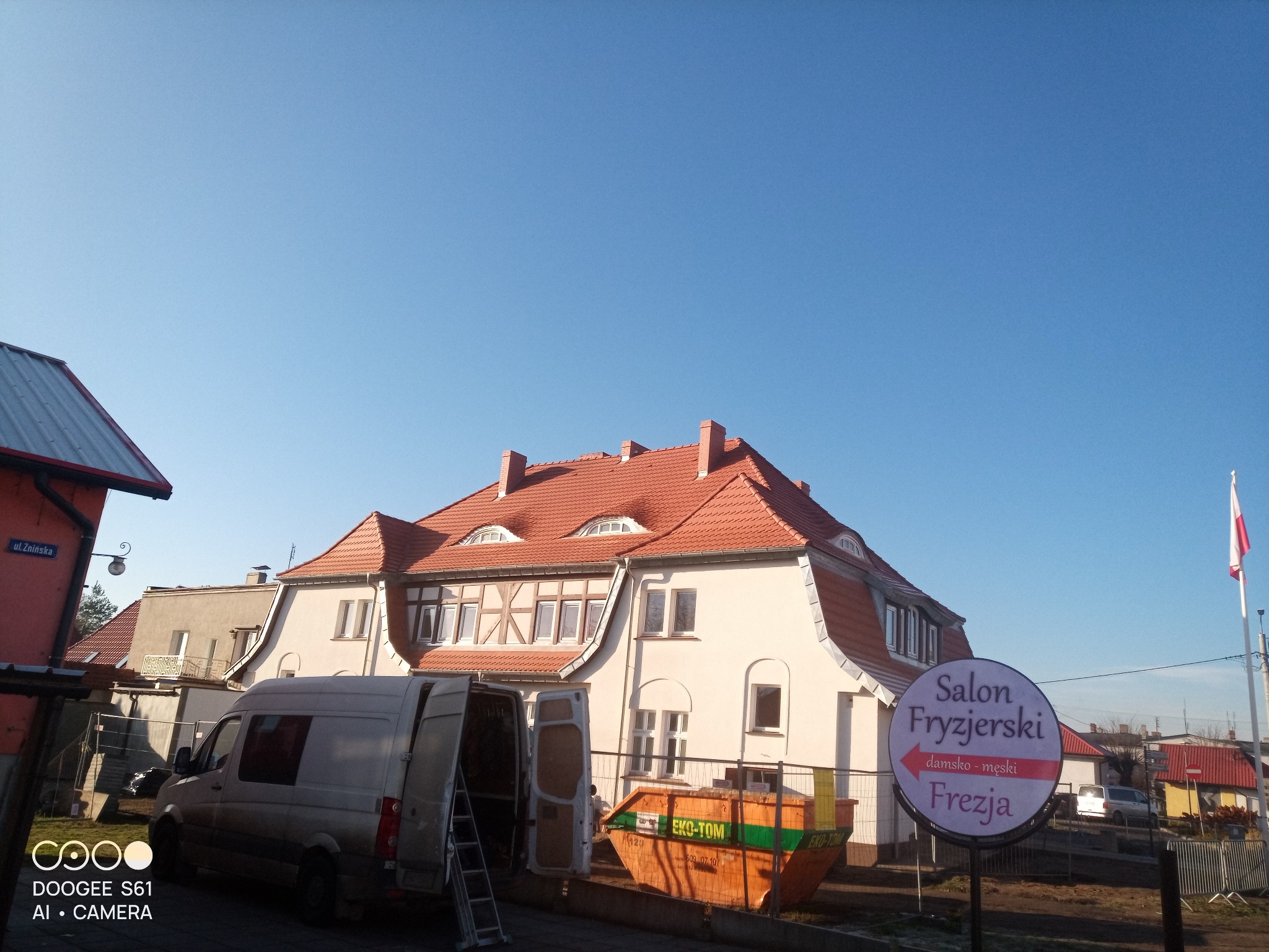 Dom z nową, czerwoną dachówką, widoczny z poziomu ulicy, z kontenerem na odpady budowlane i samochodem dostawczym z otwartymi drzwiami. Słoneczny dzień.