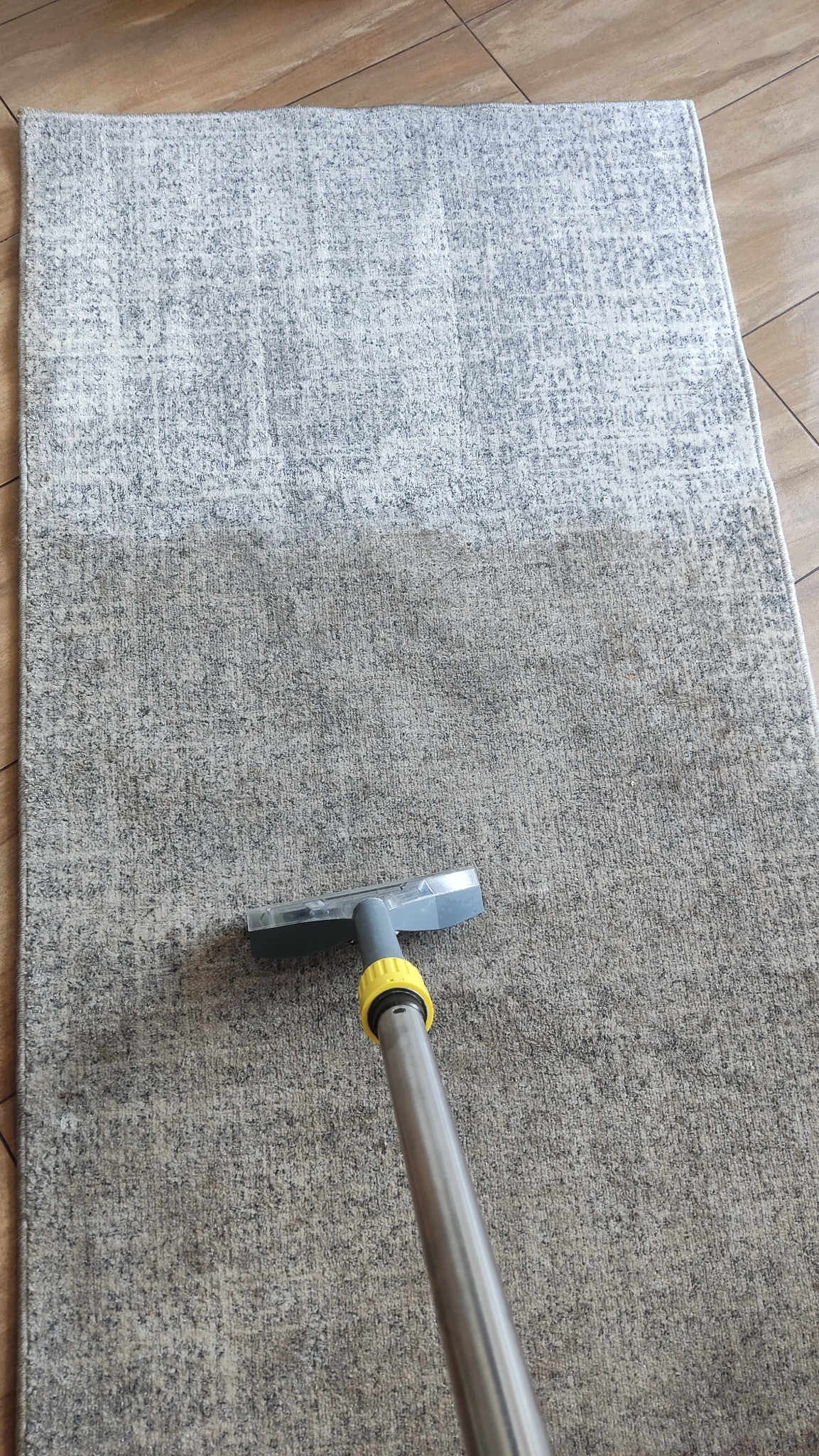 Dywan w trakcie czyszczenia odkurzaczem piorącym. Widoczna różnica w kolorze między wyczyszczoną a brudną częścią dywanu. Jasne płytki podłogowe w tle.