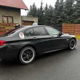 Taxi Lubań 42 - Twoje taxi - Czarna taksówka BMW z naklejką 'Lubań 42' i lampką 'TAXI' na dachu, zaparkowana na mokrej ulicy, na tle domu i zielonych drzew.