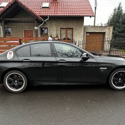 Taxi Lubań 42 - Twoje taxi - Czarna limuzyna z oznaczeniem lokalnej korporacji taksówkarskiej na tle domu z czerwoną dachówką i zielonymi drzewami. Widok z boku, mokra nawierzchnia.