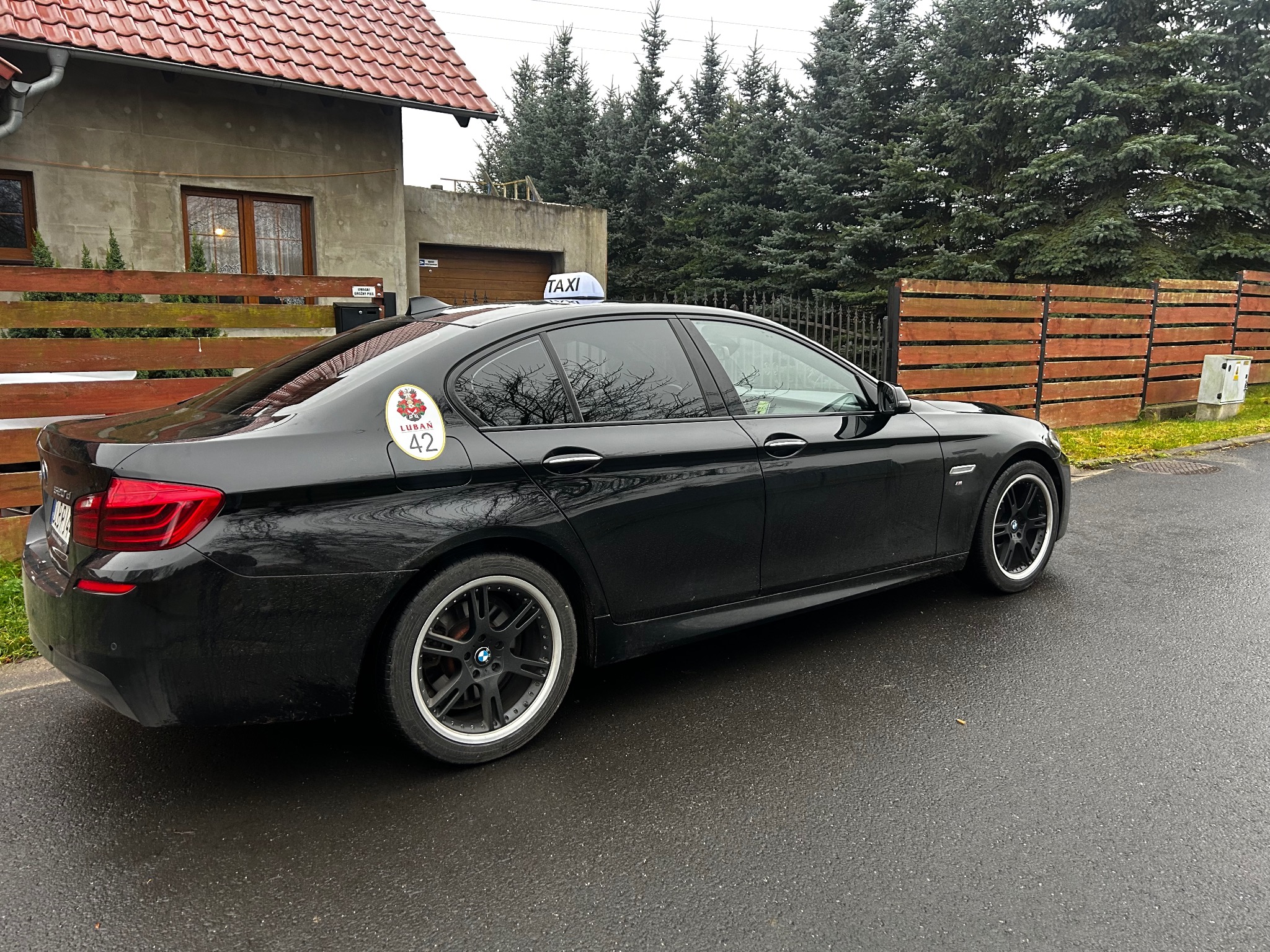 Czarna taksówka BMW z naklejką 'Lubań 42' i lampką 'TAXI' na dachu, zaparkowana na mokrej ulicy, na tle domu i zielonych drzew.