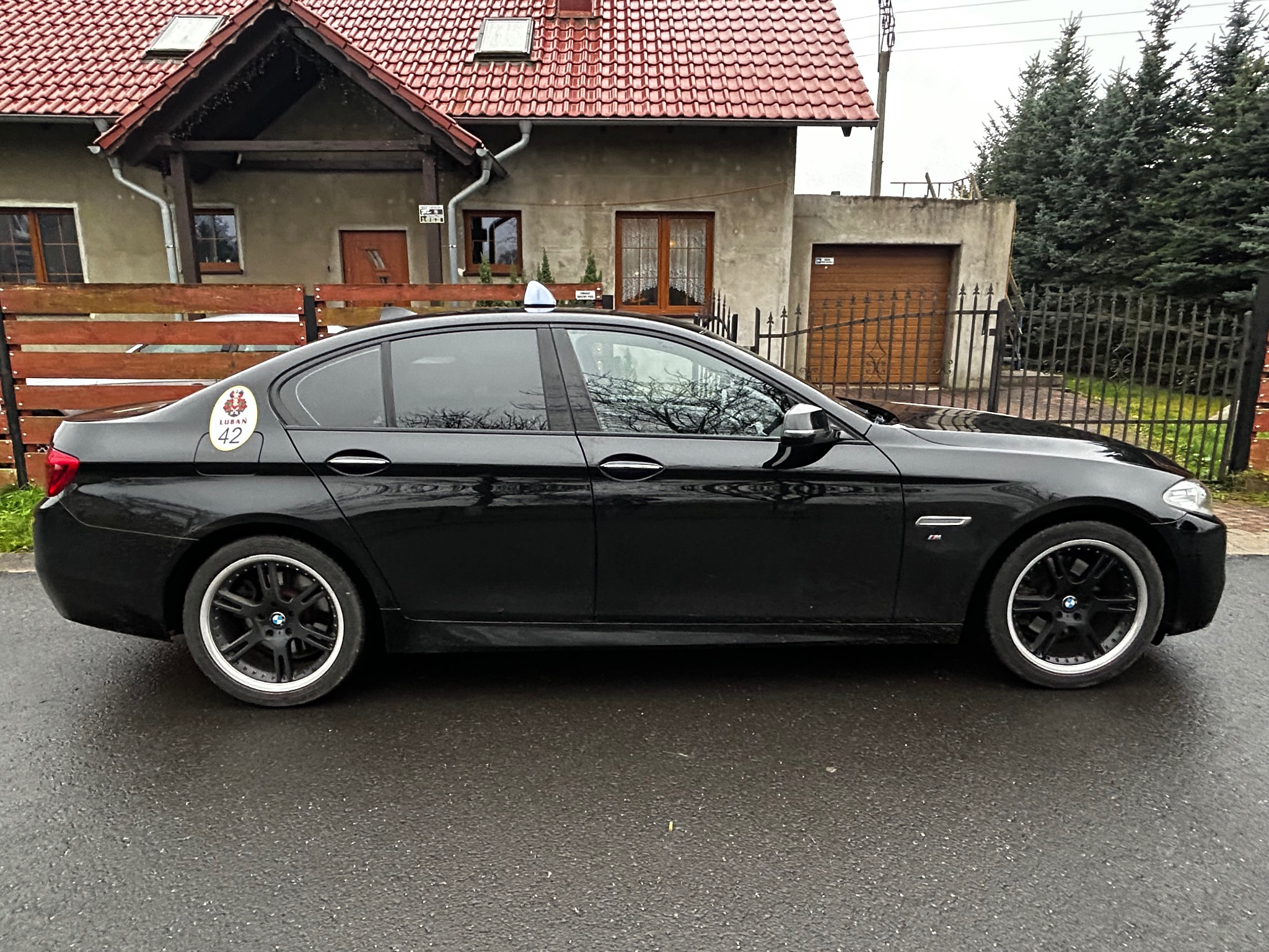 Czarna limuzyna z oznaczeniem lokalnej korporacji taksówkarskiej na tle domu z czerwoną dachówką i zielonymi drzewami. Widok z boku, mokra nawierzchnia.