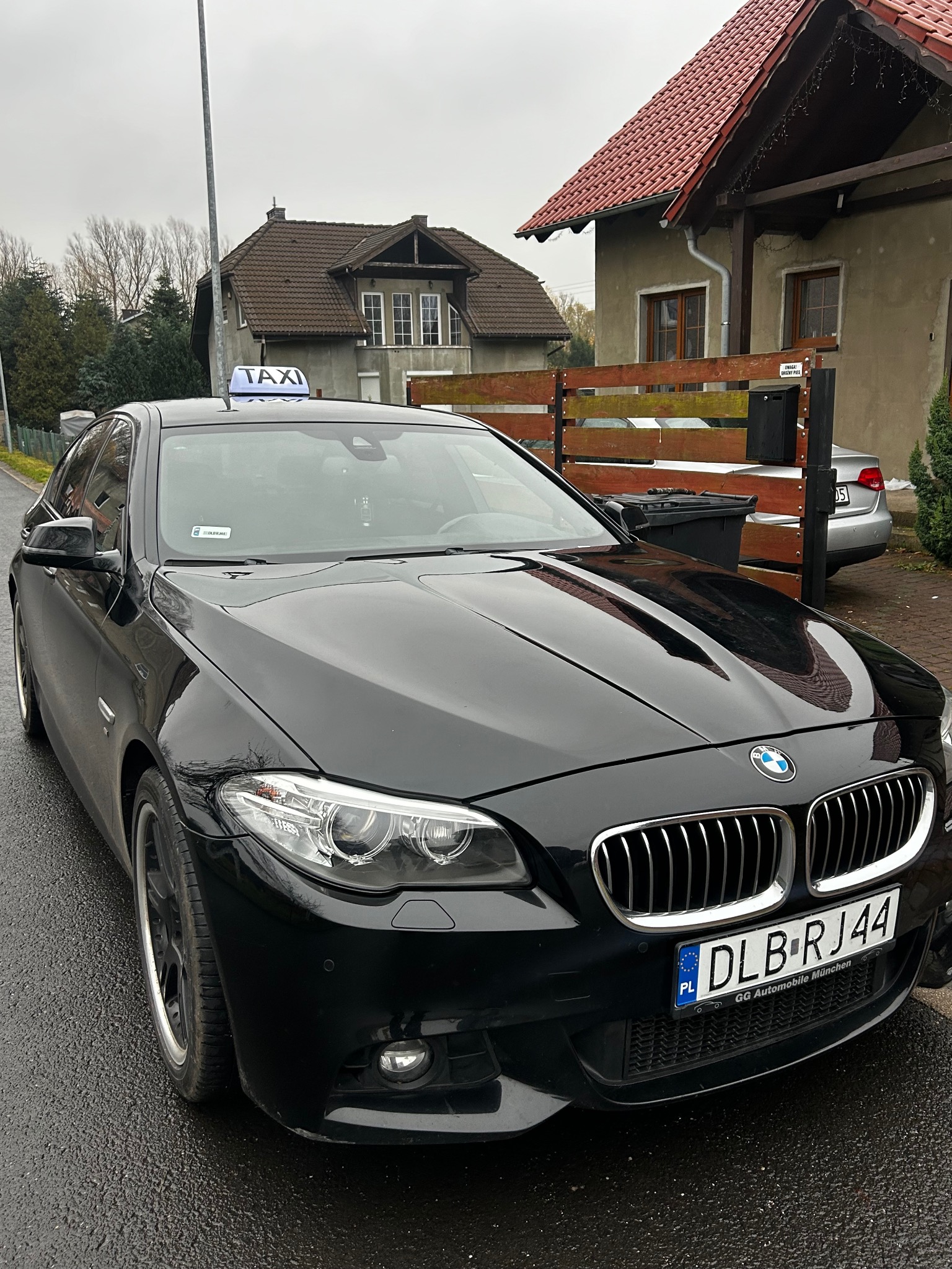 Czarna limuzyna BMW z napisem TAXI na dachu, zaparkowana na ulicy w pochmurny dzień. Widoczny numer rejestracyjny i fragment zabudowy mieszkalnej w tle.