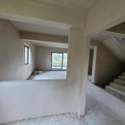 Tynki maszynowe Paweł - Wnętrze w trakcie prac wykończeniowych: gładkie, jasne ściany i sufit, widoczne schody. W tle okno z widokiem na zieleń. Na podłodze widoczne ślady prac.