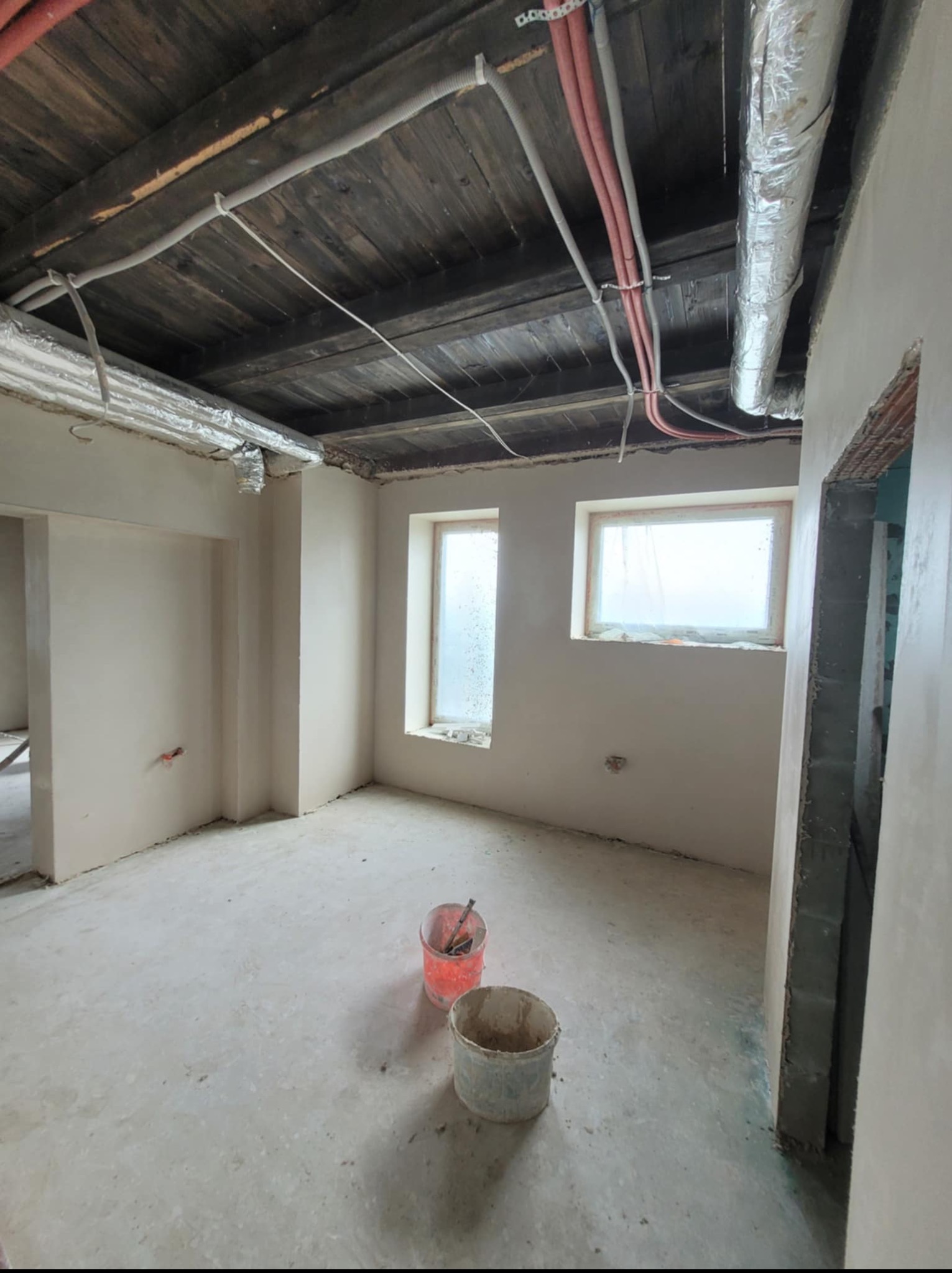 Wnętrze po nałożeniu gładzi, widoczne dwa wiadra na podłodze, instalacje pod sufitem i okna. Ściany gładkie, przygotowane do malowania. Surowy stan wykończenia.