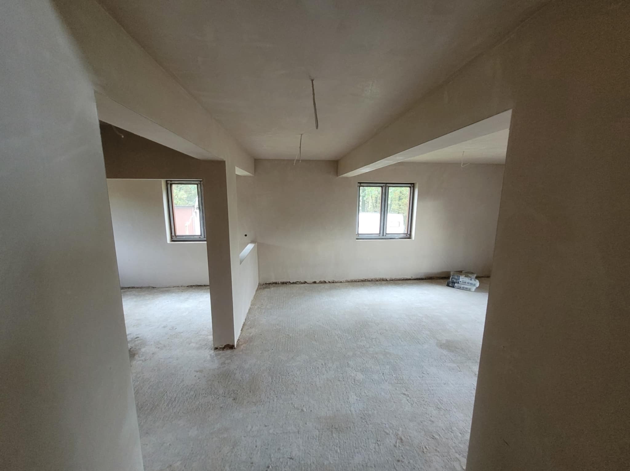 Wnętrze po nałożeniu gładzi: jasne ściany, surowy beton na podłodze, okna z widokiem na zieleń. Worki z materiałem budowlanym w rogu pomieszczenia.