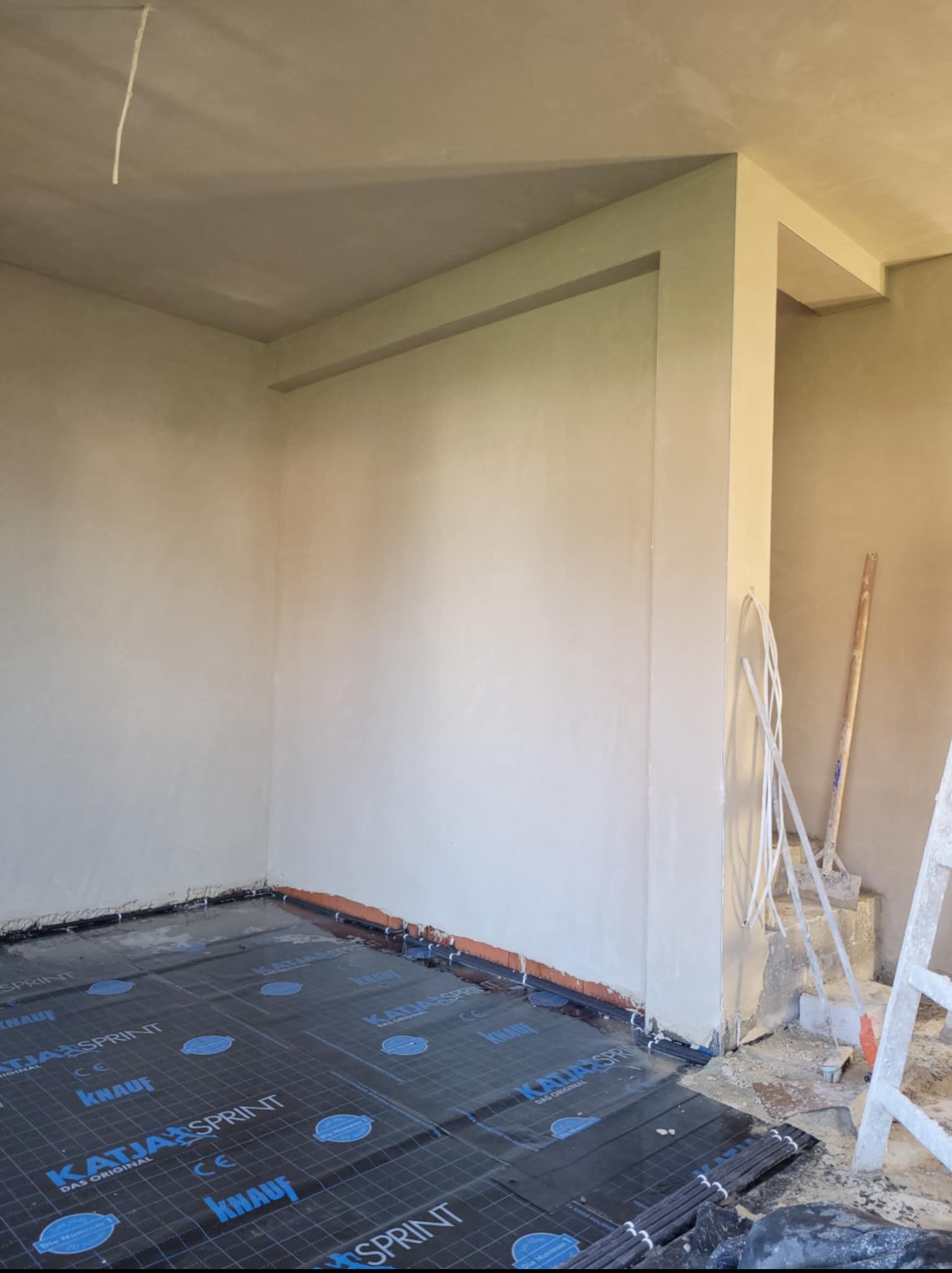 Wnętrze po tynkowaniu maszynowym: gładkie, jasne ściany, folia izolacyjna na podłodze, widoczne narzędzia i drabina. Przygotowanie do dalszych prac wykończeniowych.