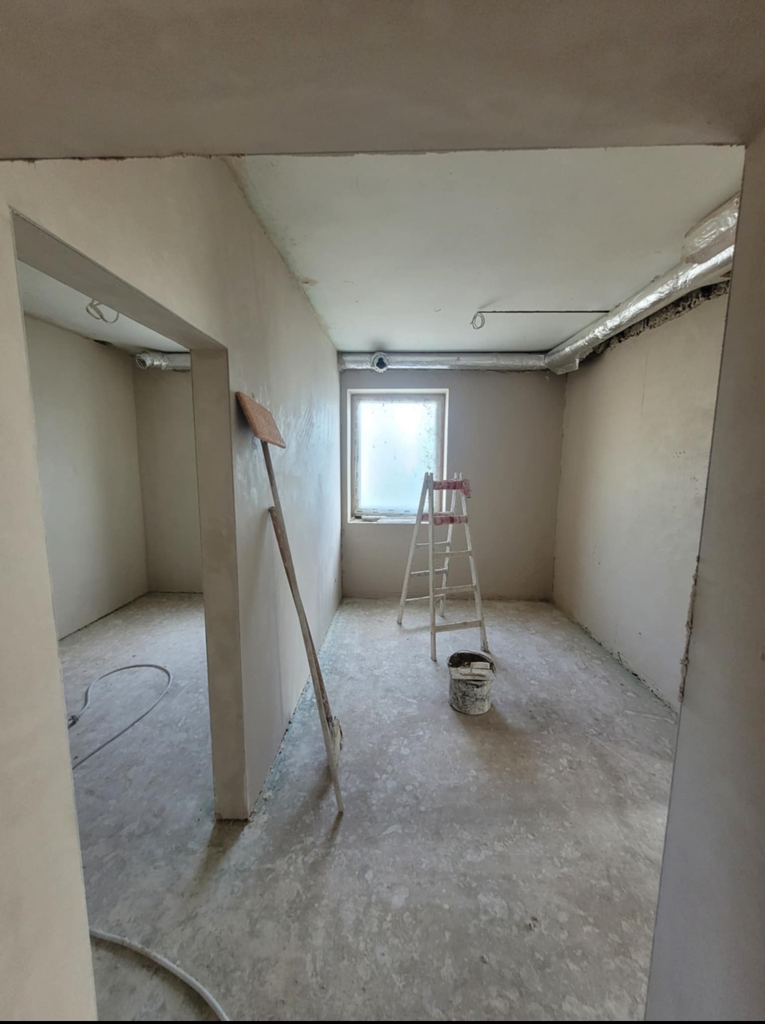 Pokój po nałożeniu gładzi, widoczna drabina, wiadro i narzędzie oparta o ścianę. Ściany i sufit pokryte świeżą, jasną gładzią. Widok z perspektywy wejścia.
