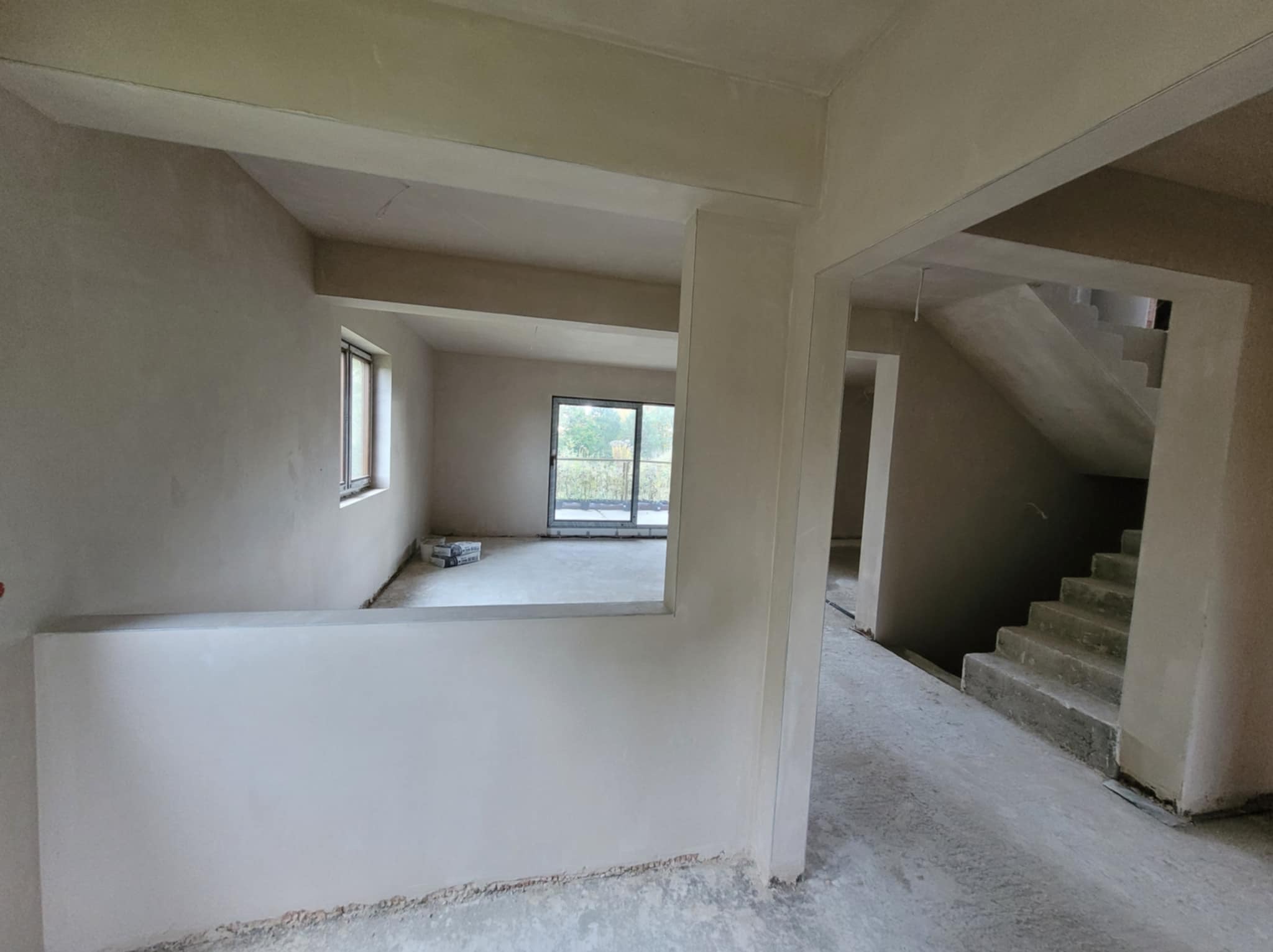 Wnętrze w trakcie prac wykończeniowych: gładkie, jasne ściany i sufit, widoczne schody. W tle okno z widokiem na zieleń. Na podłodze widoczne ślady prac.