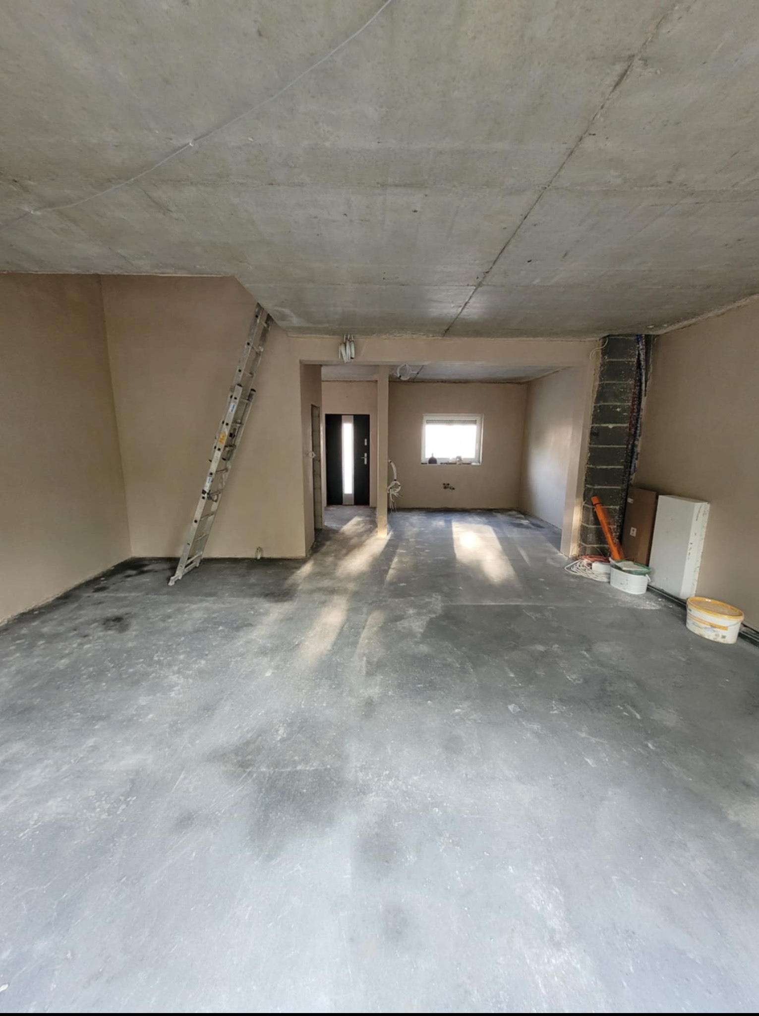 Wnętrze w trakcie prac wykończeniowych: ściany po szpachlowaniu, surowy betonowy sufit, drabina oparta o ścianę, widoczne puszki z farbą i narzędzia.