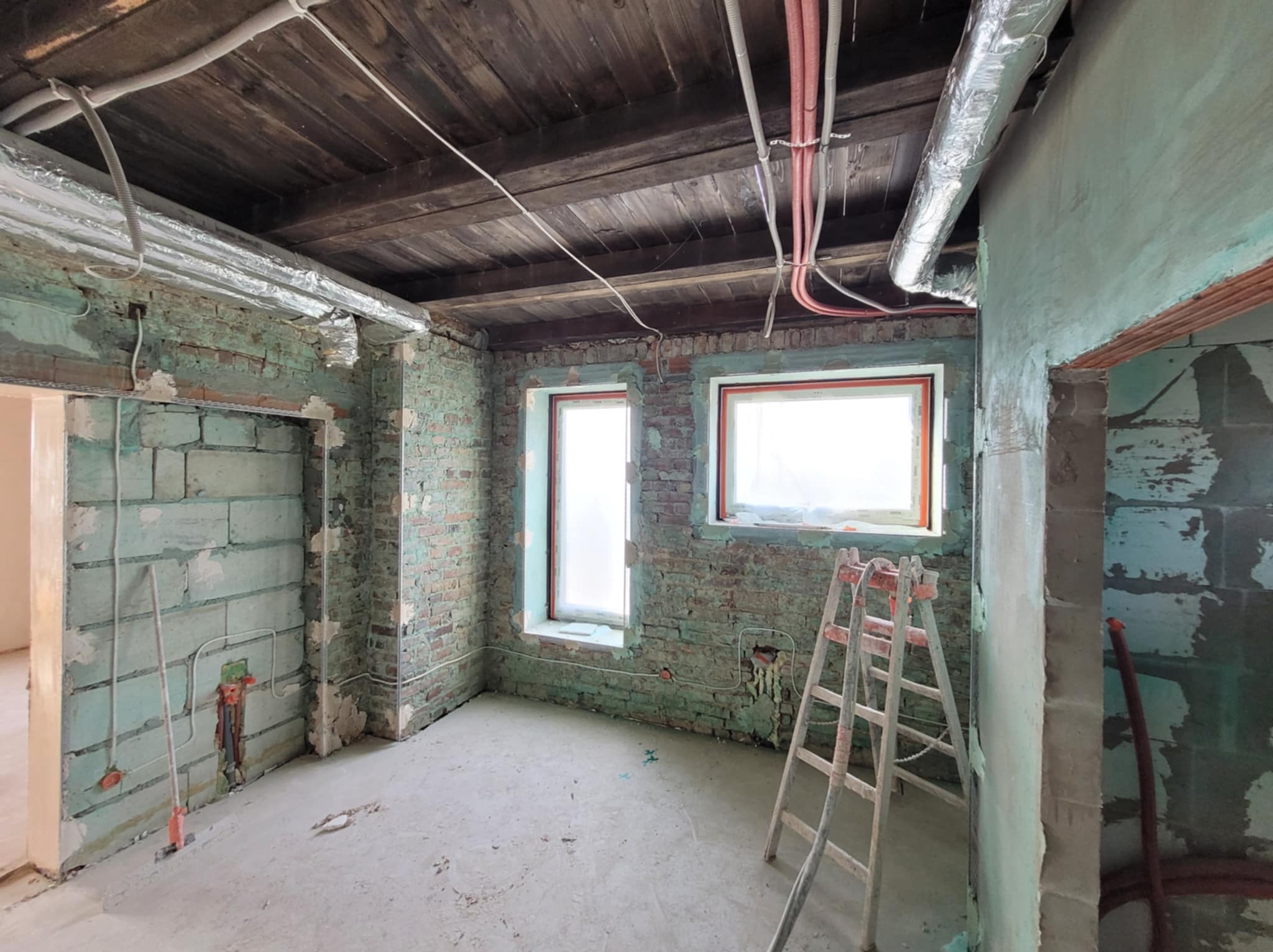 Wnętrze w trakcie remontu: surowe ściany z instalacjami, drewniany sufit z rurami wentylacyjnymi, drabina oparta o ścianę. Widoczne okna z folią ochronną.
