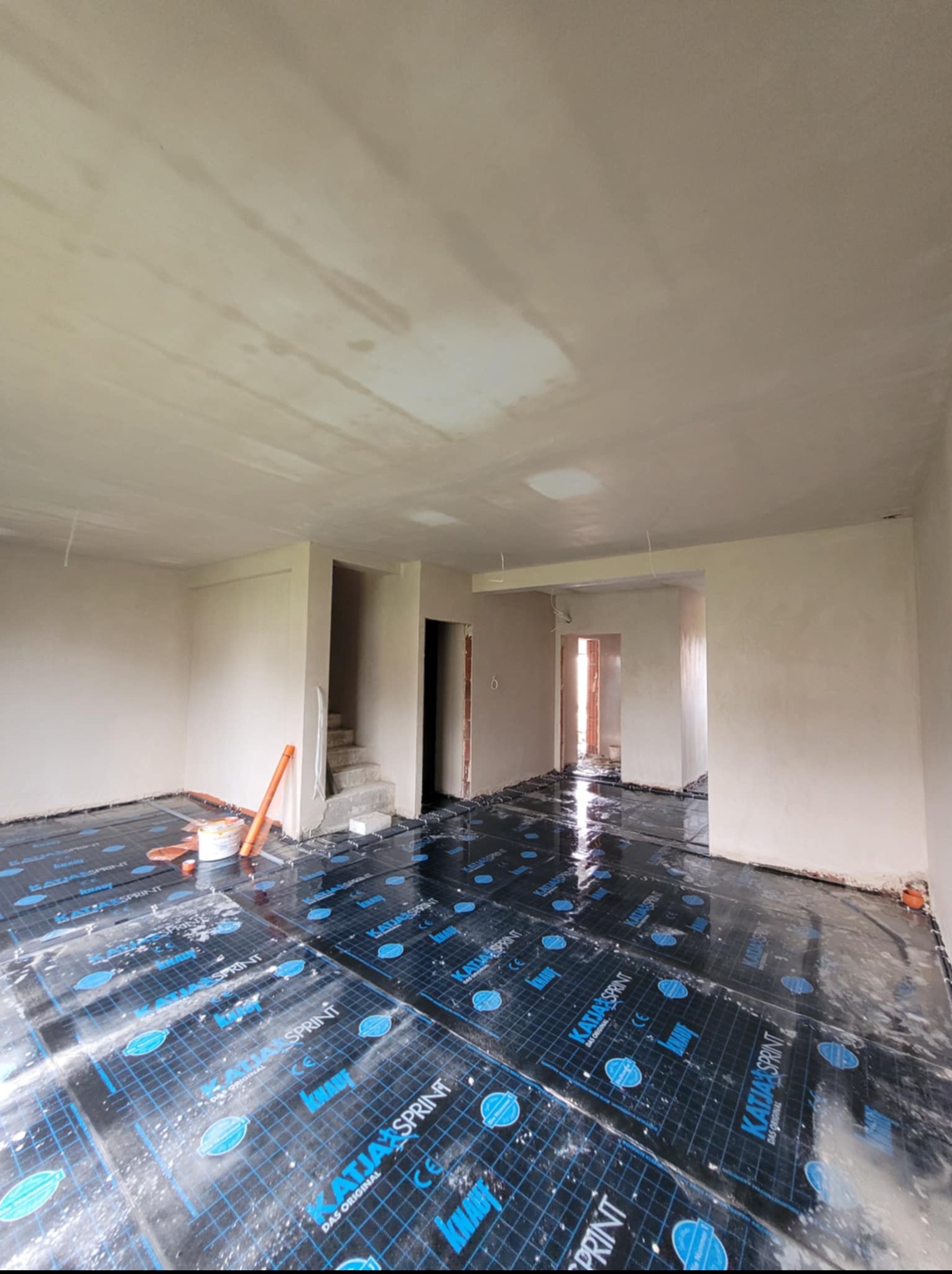 Wnętrze w trakcie prac wykończeniowych: świeżo położona folia grzewcza na podłodze, ściany i sufit po szpachlowaniu, widoczne przejścia do innych pomieszczeń.