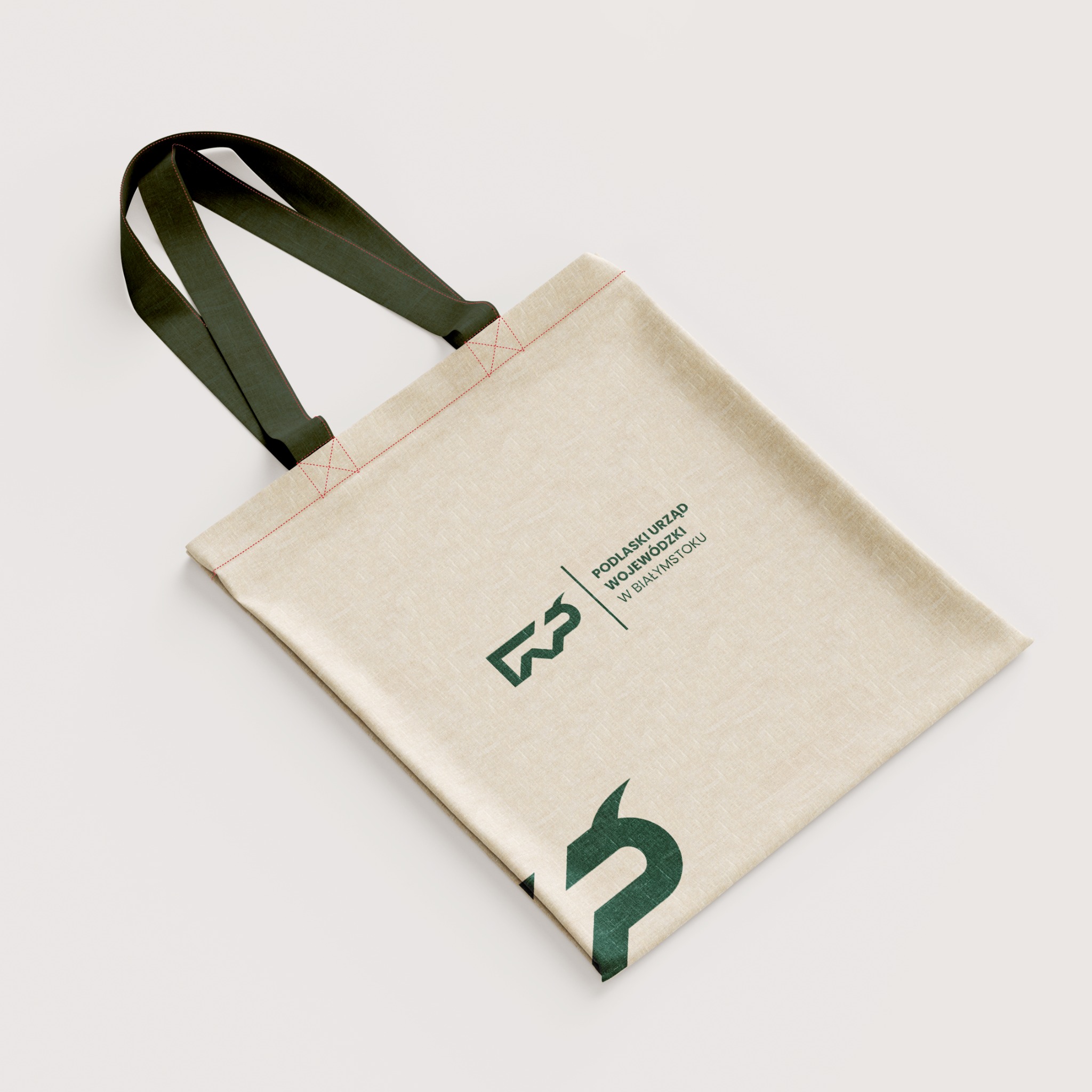 Beżowa torba bawełniana z zielonymi uchwytami i nadrukiem logo Podlaskiego Urzędu Wojewódzkiego w Białymstoku, leżąca ukośnie na białym tle.