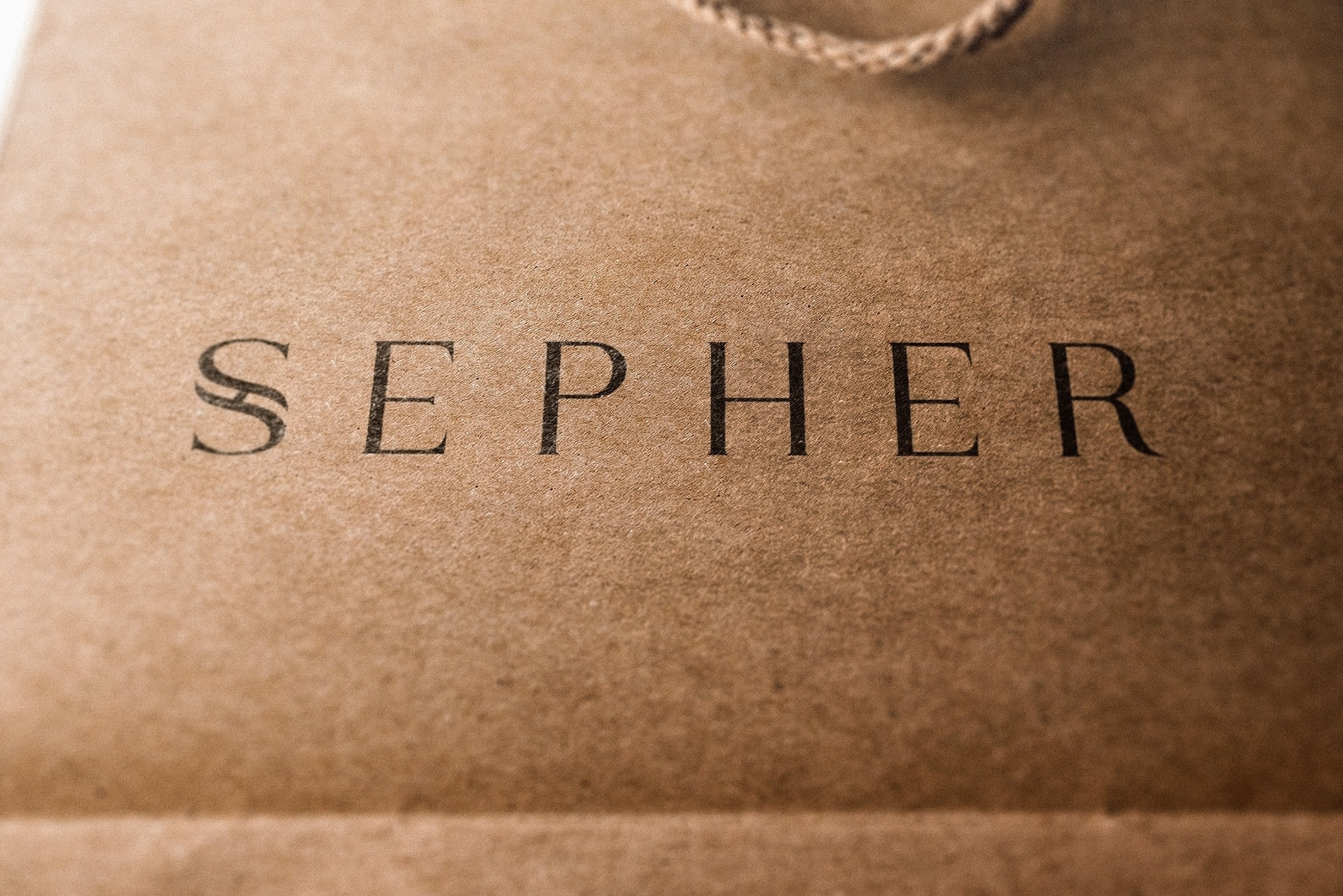 Brązowa torba papierowa z czarnym napisem 'SEPHER' w zbliżeniu, widoczny fragment sznurkowego uchwytu. Minimalistyczny design, ciepłe oświetlenie.