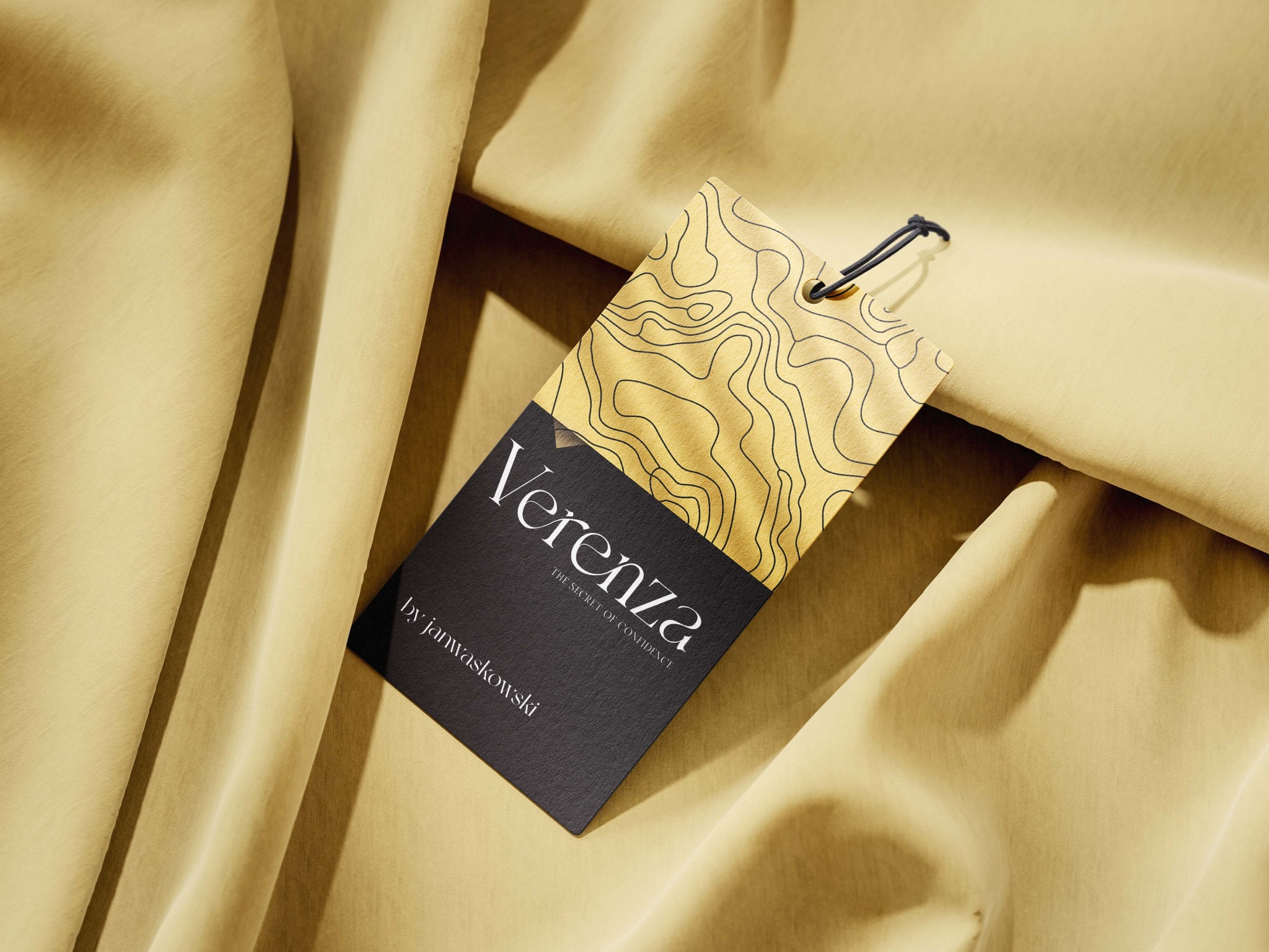 Elegancka metka z logo 'Verenza' na beżowym tle materiału. Górna część metki w kolorze złota z czarnym wzorem topograficznym, dolna czarna z białym napisem 'by janwaskowski'.