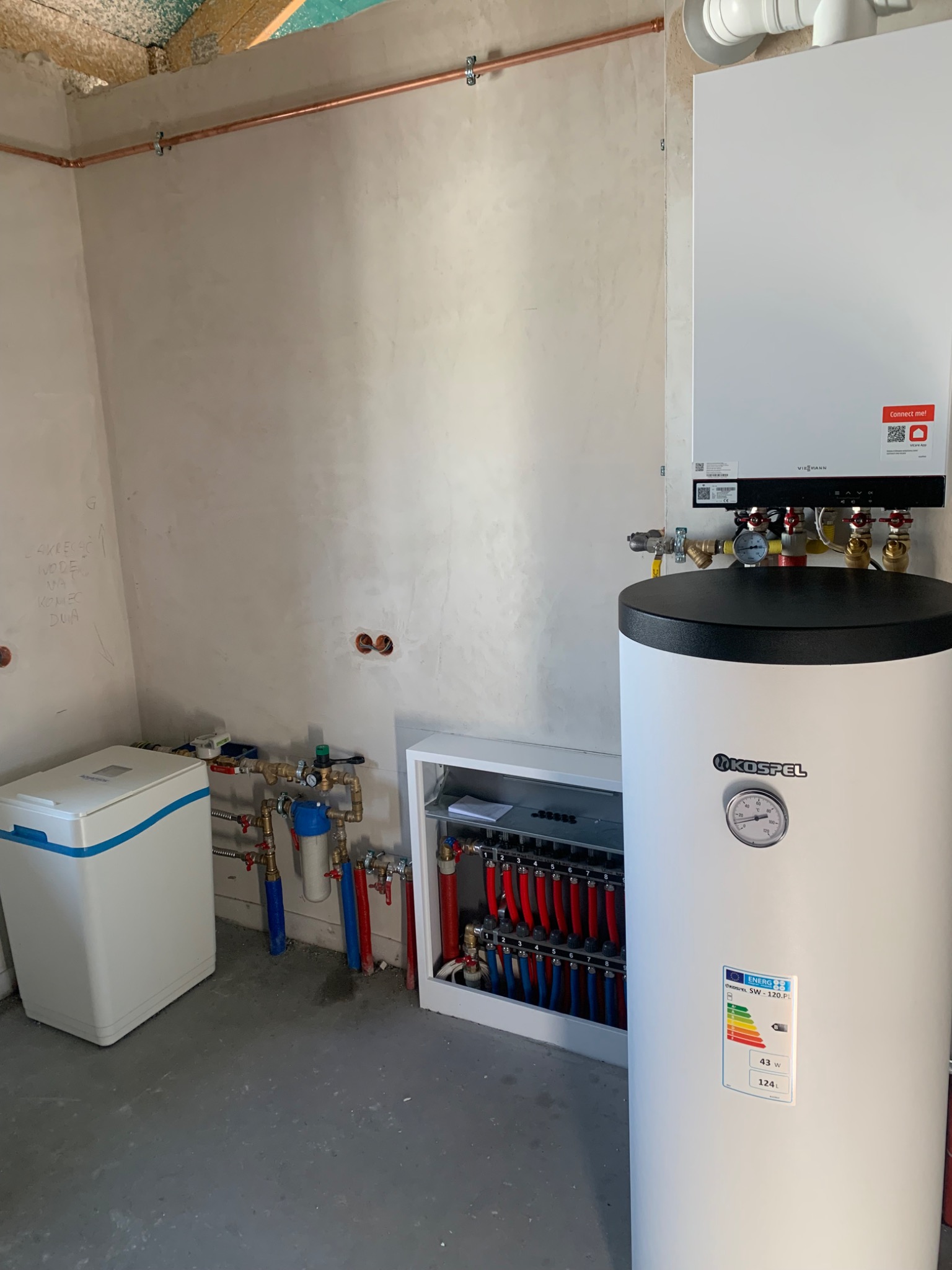 Nowoczesna kotłownia: biały zasobnik Kospel, piec Viessmann, rozdzielacz ogrzewania podłogowego i zmiękczacz wody. Miedziane rury na ścianie. Surowy tynk.