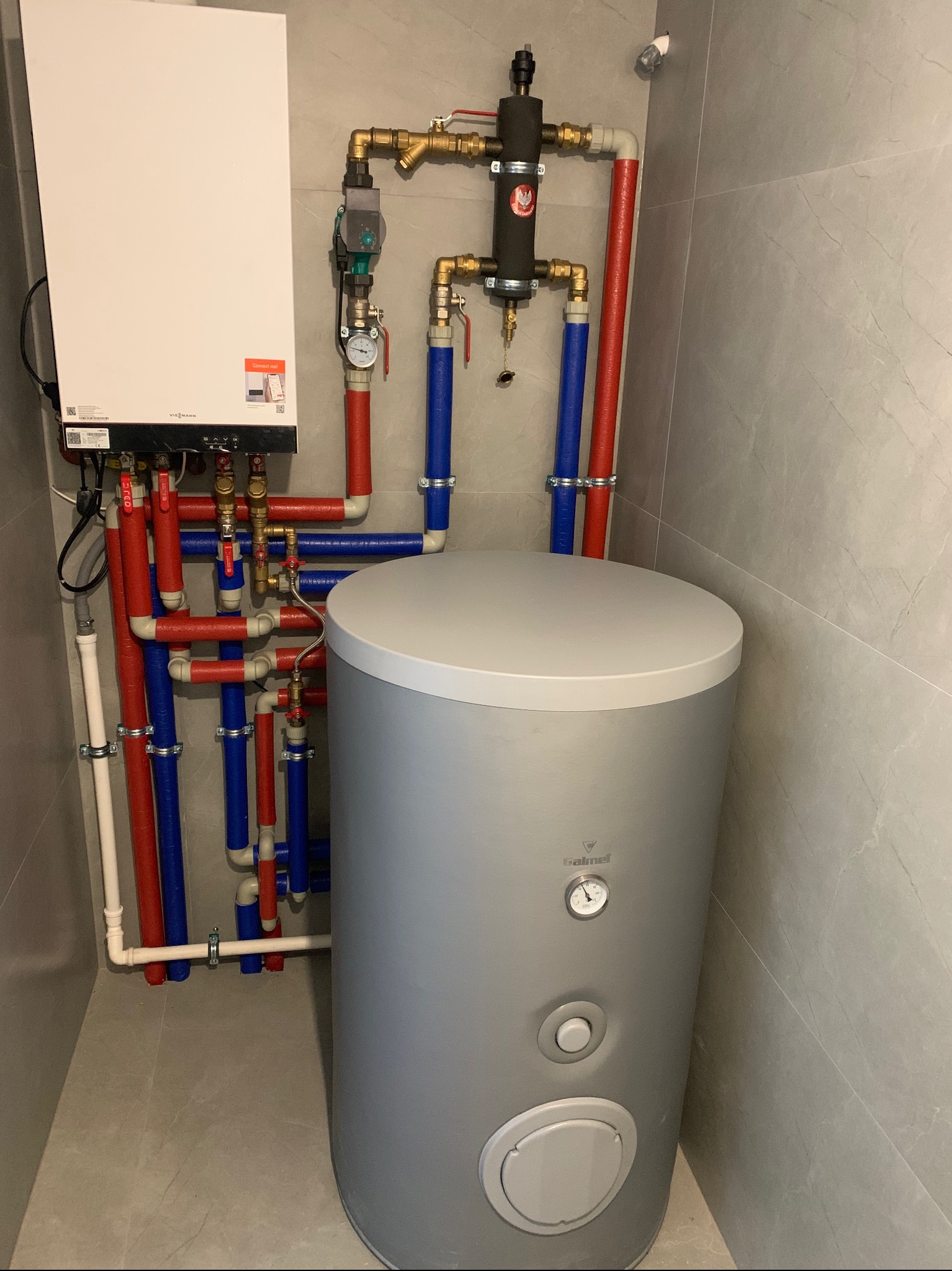 Nowoczesna kotłownia z białym piecem Viessmann i szarym zasobnikiem Galmet. Instalacja z rur w izolacji czerwonej i niebieskiej na tle szarych płytek.