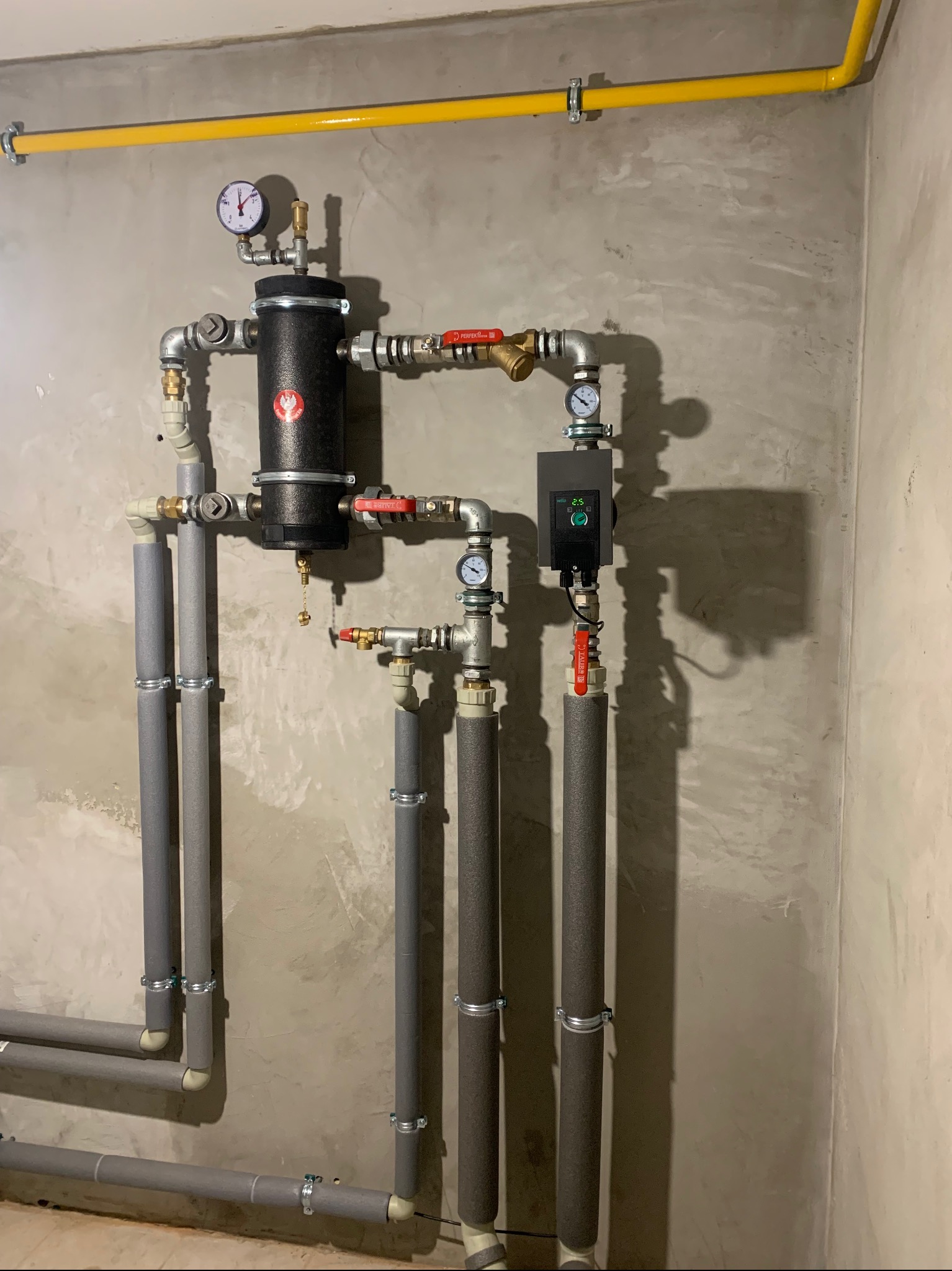 Złożona instalacja grzewcza z czarnym zbiornikiem, manometrami i rurami w izolacji na tle surowej ściany. Żółta rura gazowa u góry. Elementy instalacji połączone zaworami.