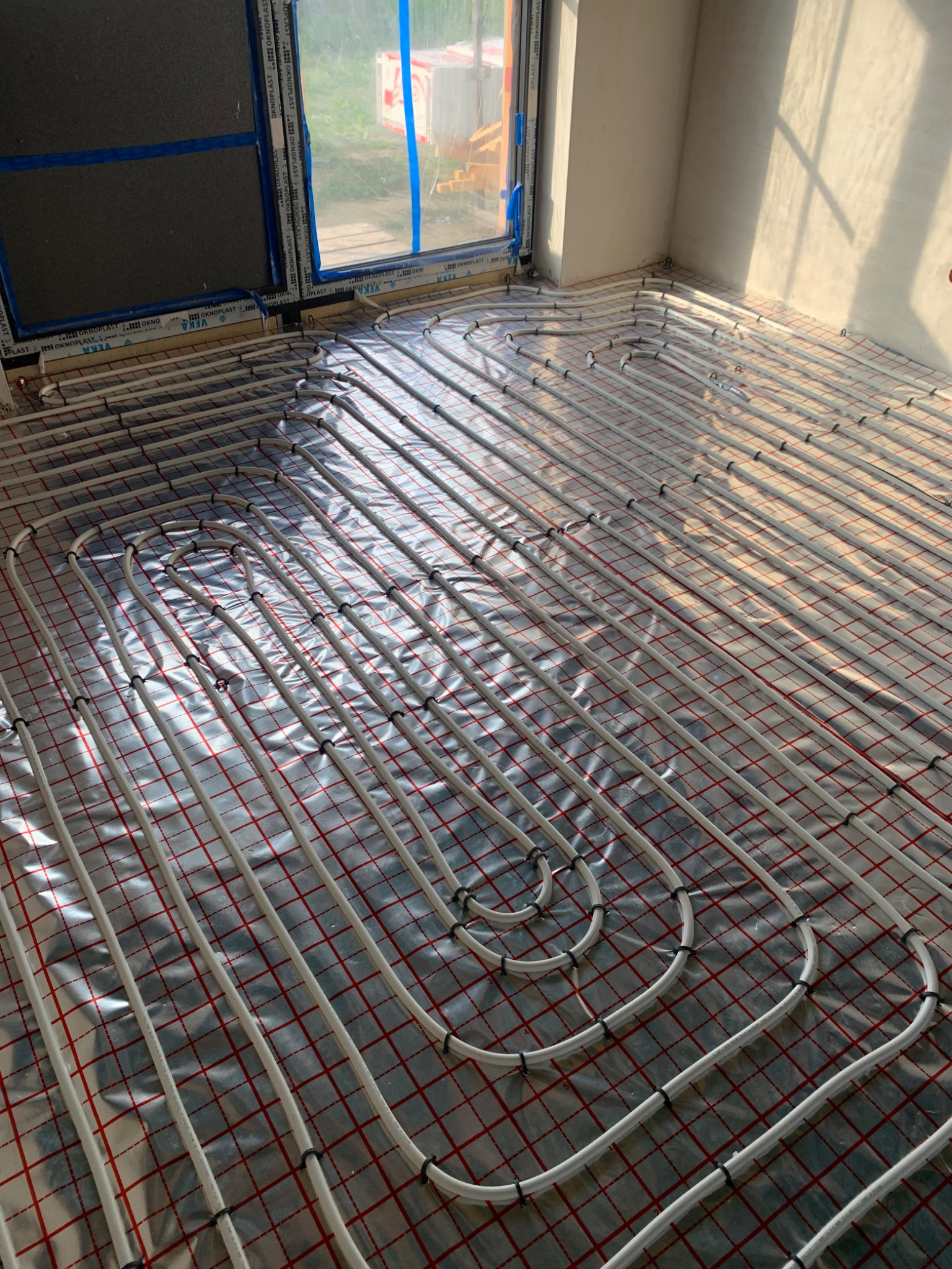 Białe rury ogrzewania podłogowego, ułożone na folii z czerwoną siatką, w jasnym pomieszczeniu z oknem. Widoczne mocowania rur. Etap instalacji przed wylewką.