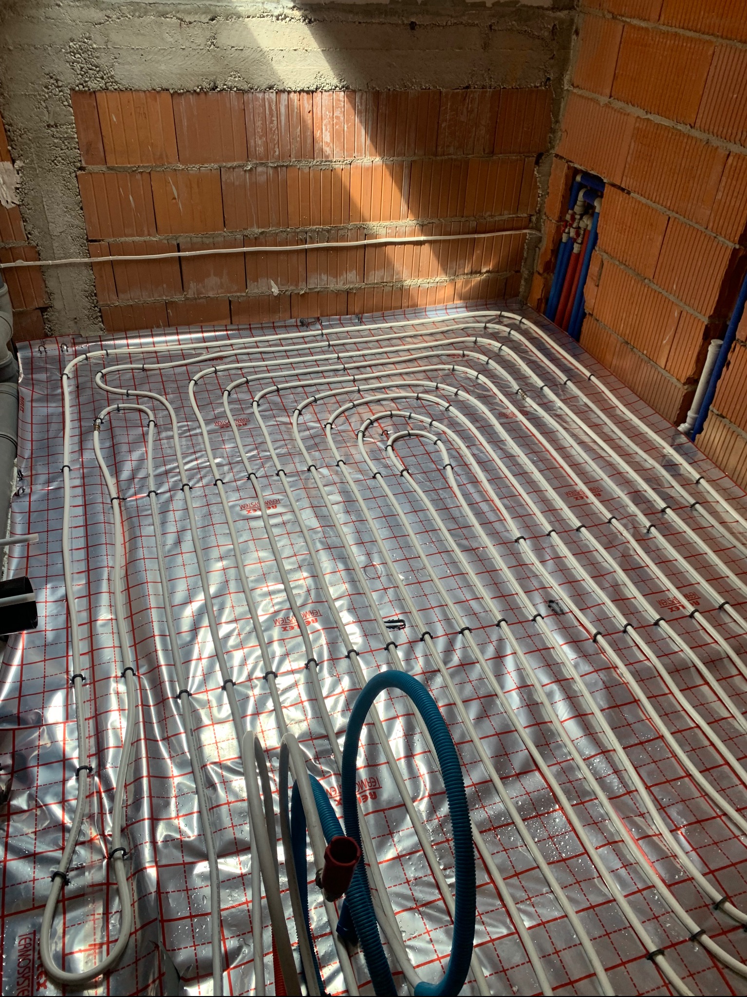Białe rury ogrzewania podłogowego ułożone na srebrnej folii z czerwoną siatką, widoczne fragmenty ściany z czerwonej cegły i niebieskie rury instalacji.