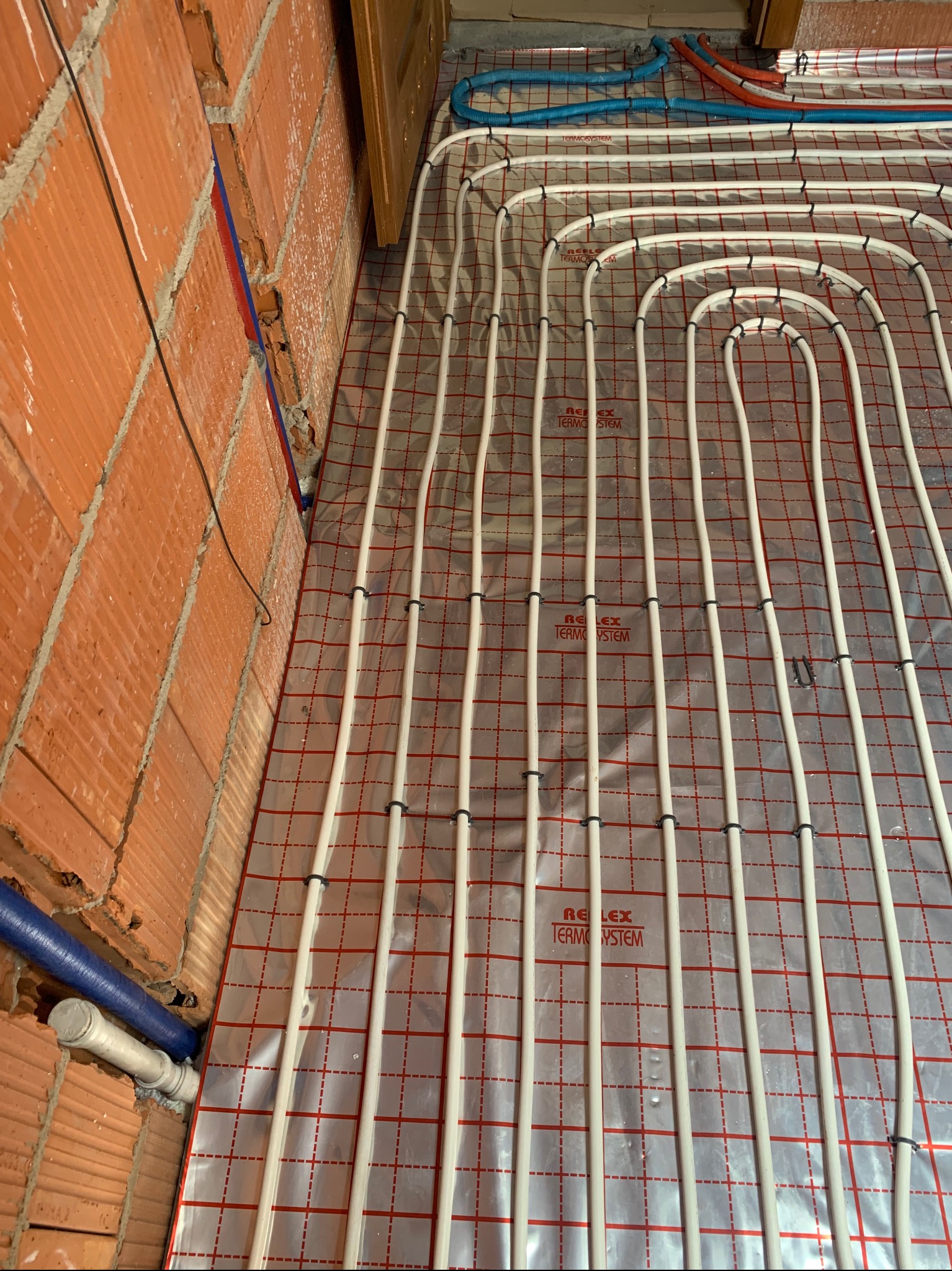 Białe rury ogrzewania podłogowego ułożone na folii z czerwoną siatką, widoczne mocowania i fragment ściany z cegły. Instalacja w trakcie realizacji.