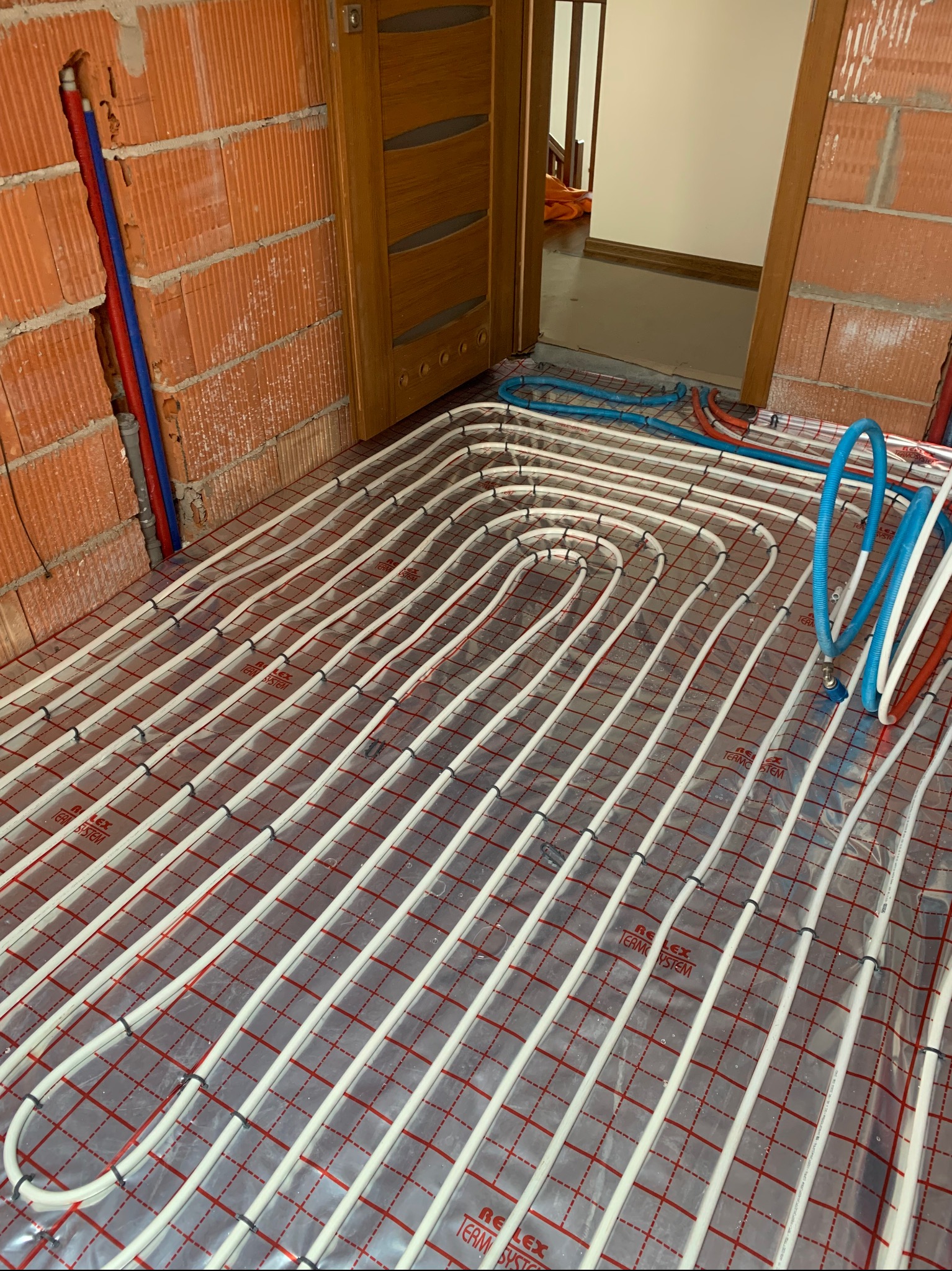 Instalacja ogrzewania podłogowego w trakcie montażu. Białe rurki ułożone na folii z czerwoną siatką, widoczne przyłącza do ściany z cegły dziurawki i fragment drzwi.