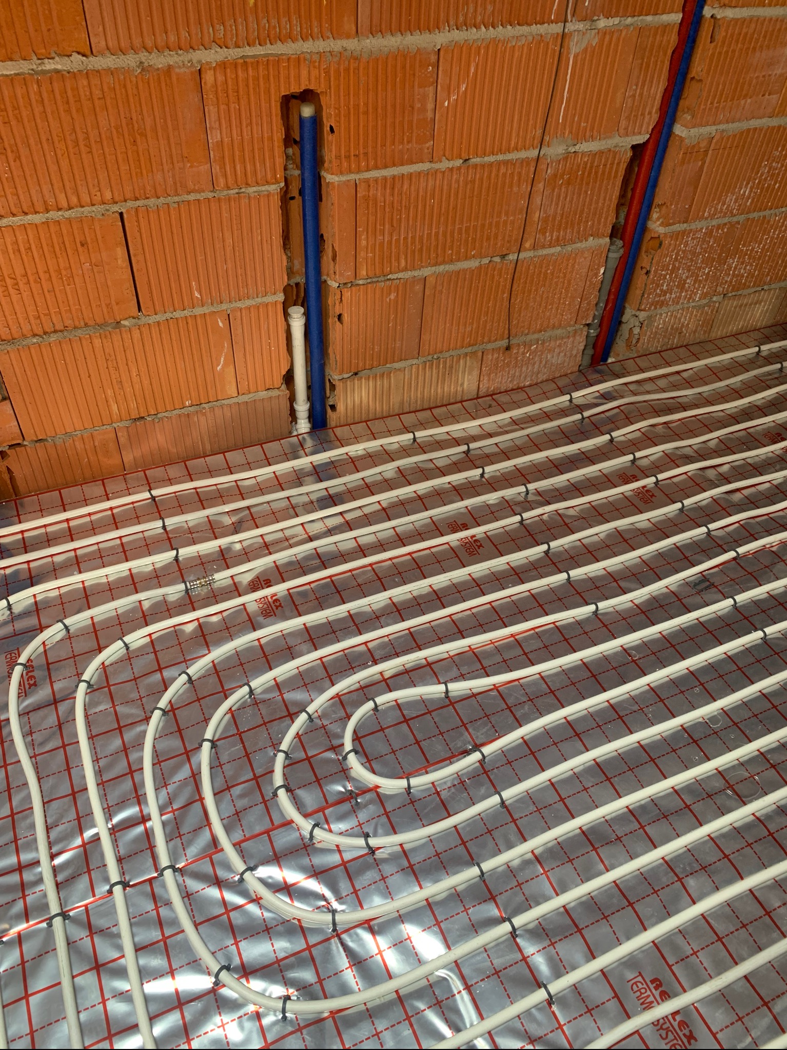 Białe rurki ogrzewania podłogowego ułożone na folii z czerwoną siatką, widoczne pionowe rury w ścianie z cegły dziurawki. Instalacja w trakcie montażu.
