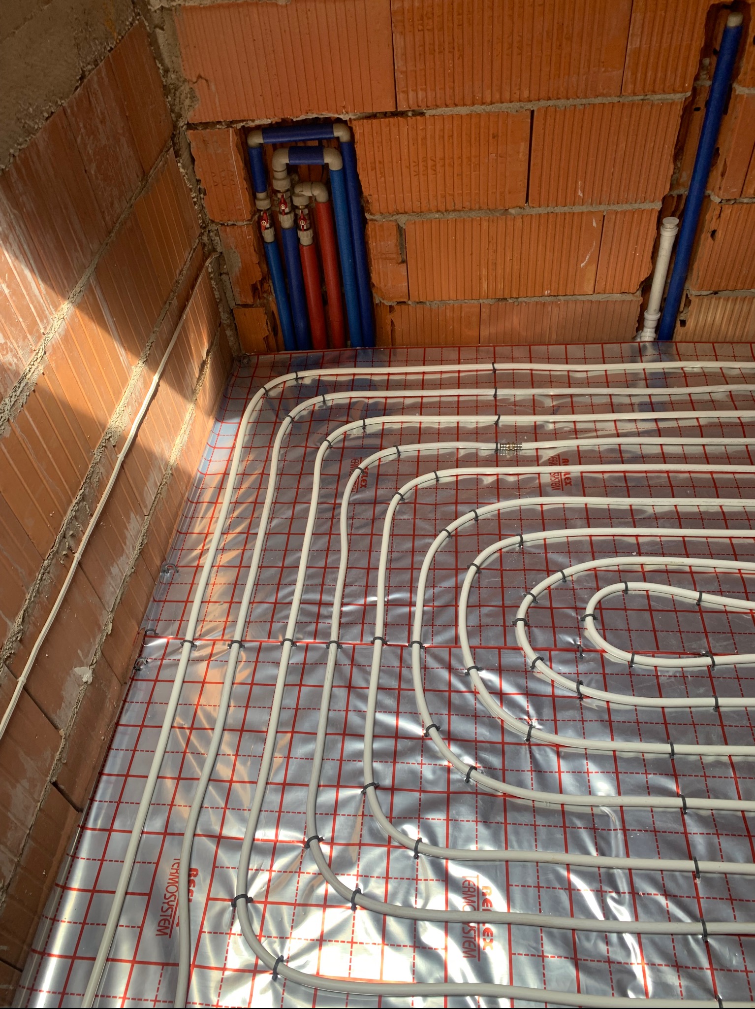 Instalacja ogrzewania podłogowego w surowym wnętrzu z ceglanymi ścianami. Białe rurki ułożone na folii z czerwoną siatką, widoczne niebieskie i czerwone rury w ścianie.