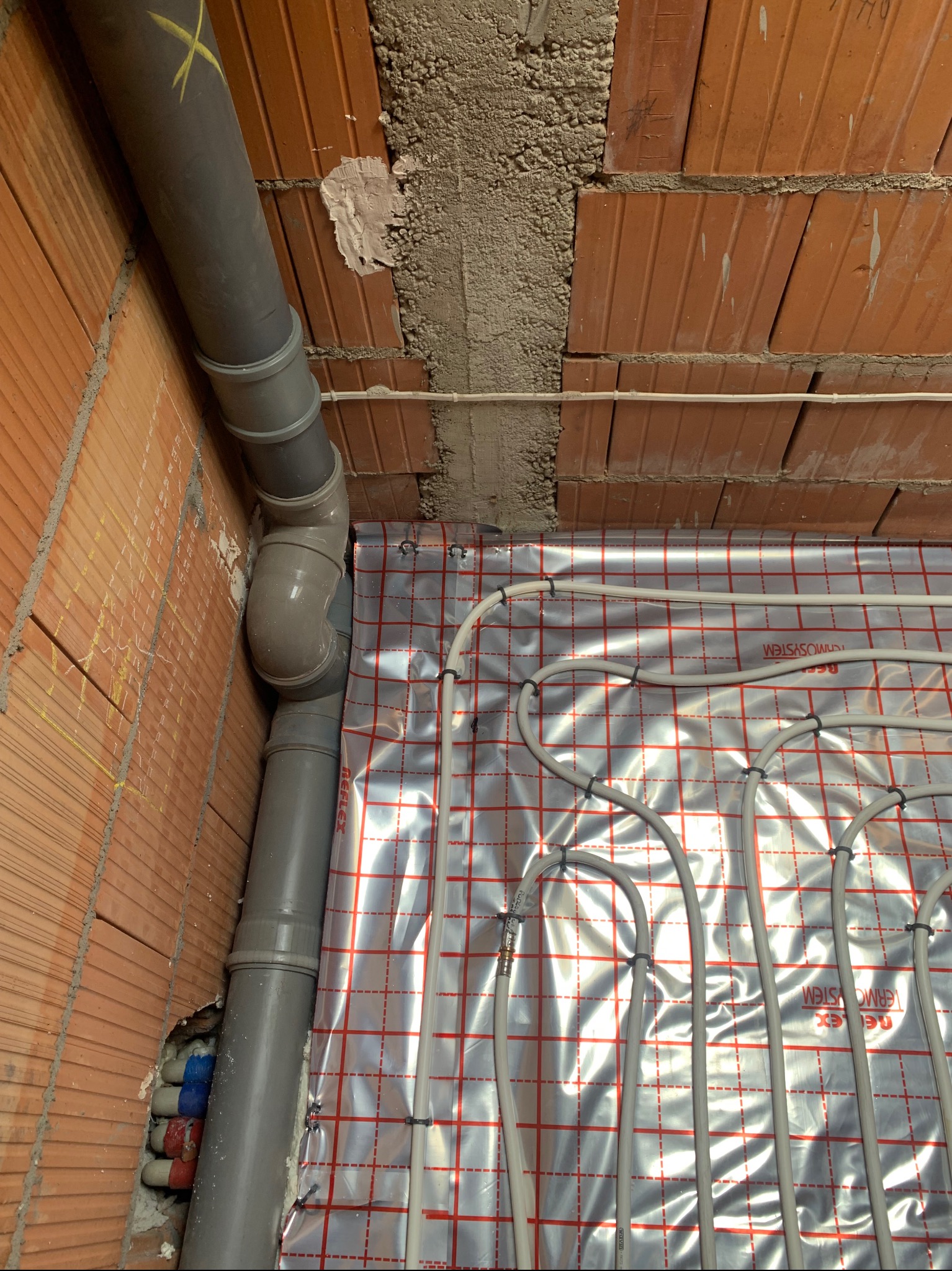 Ułożone rury ogrzewania podłogowego na folii refleksyjnej, widoczne pionowe rury kanalizacyjne przy ceglanej ścianie. Etap instalacji przed wylewką.