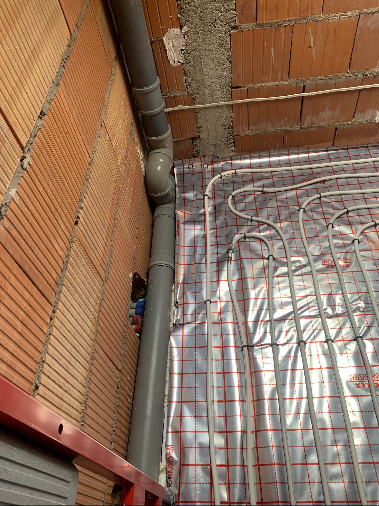 Instalacja ogrzewania podłogowego w trakcie montażu: rury grzewcze ułożone na folii refleksyjnej, widoczne przyłącza i fragment ściany z cegły.